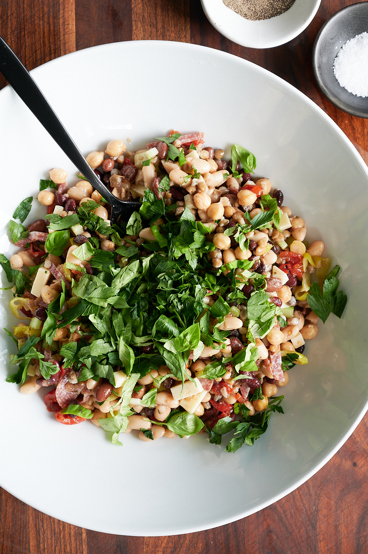A large white bowl filled with mixed bean salad topped with chopped fresh herbs, with a black serving spoon and bowls of salt and pepper nearby.