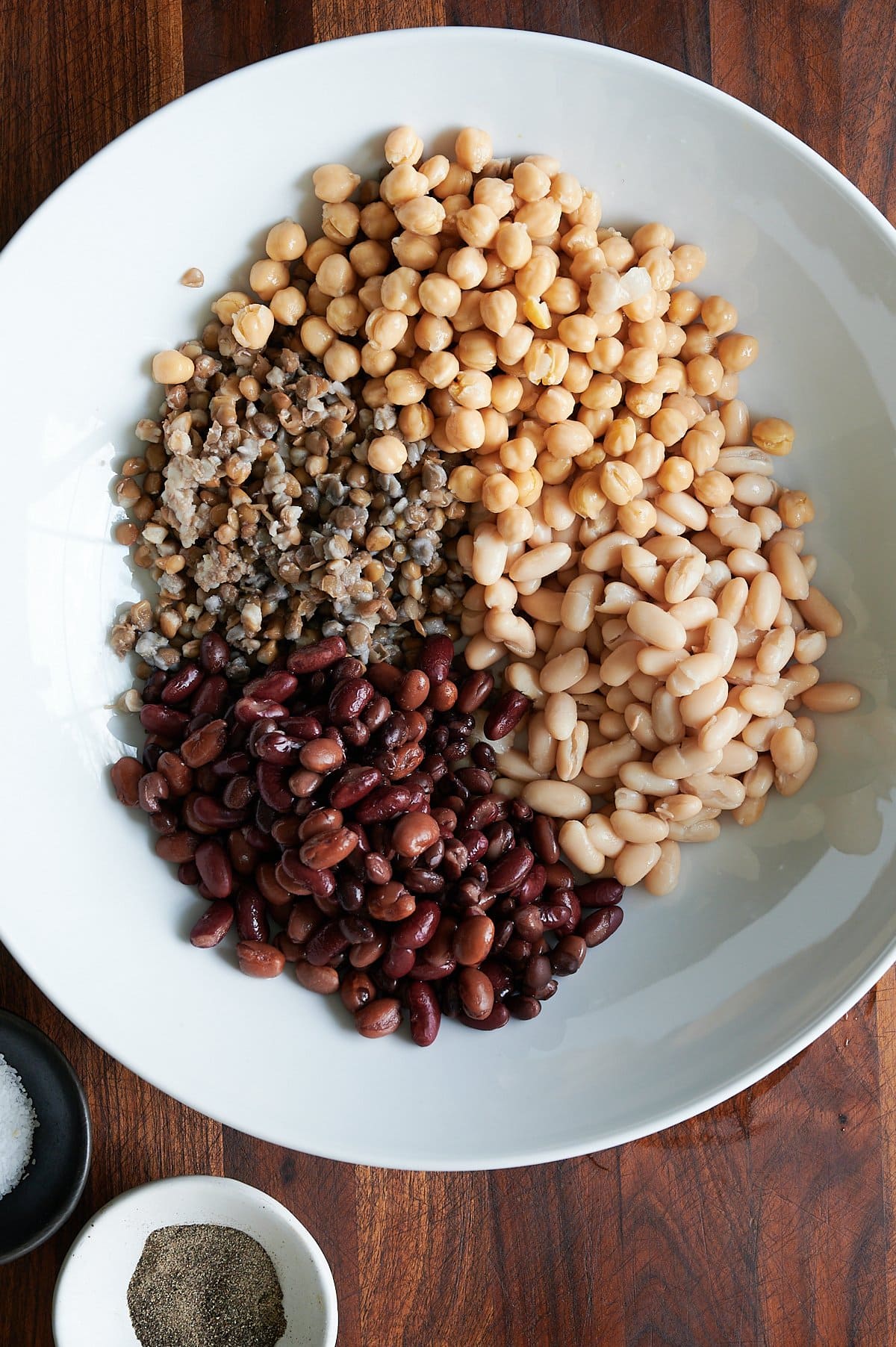 A white bowl with chickpeas, white beans, kidney beans, and lentils, next to small bowls of salt and pepper on a wooden surface.