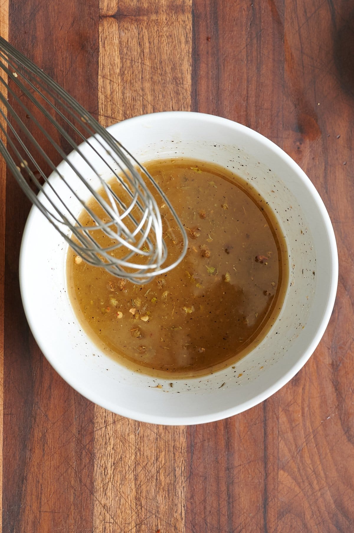 A metal whisk rests in a white bowl of salad dressing on a wooden cutting board.