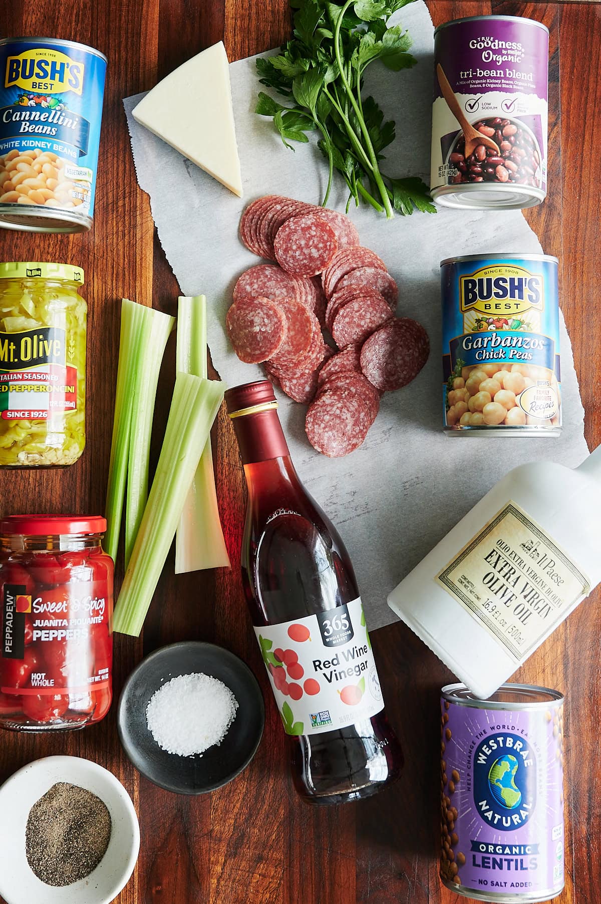 A top-down view of bean salad ingredients on a wooden surface, including deli meats, cheeses, canned beans, herbs, celery, oil, vinegar, salt, pepper, and peppers.