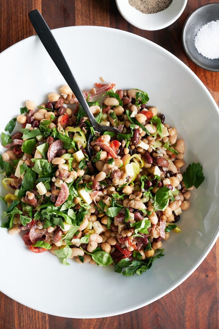 A white bowl filled with dense bean salad, herbs, chopped vegetables, and a black serving spoon, placed on a wooden surface next to bowls of salt and pepper.