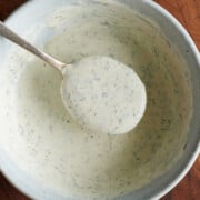 A spoon holding up creamy, herb-speckled ranch dressing over a light gray bowl on a wooden surface.