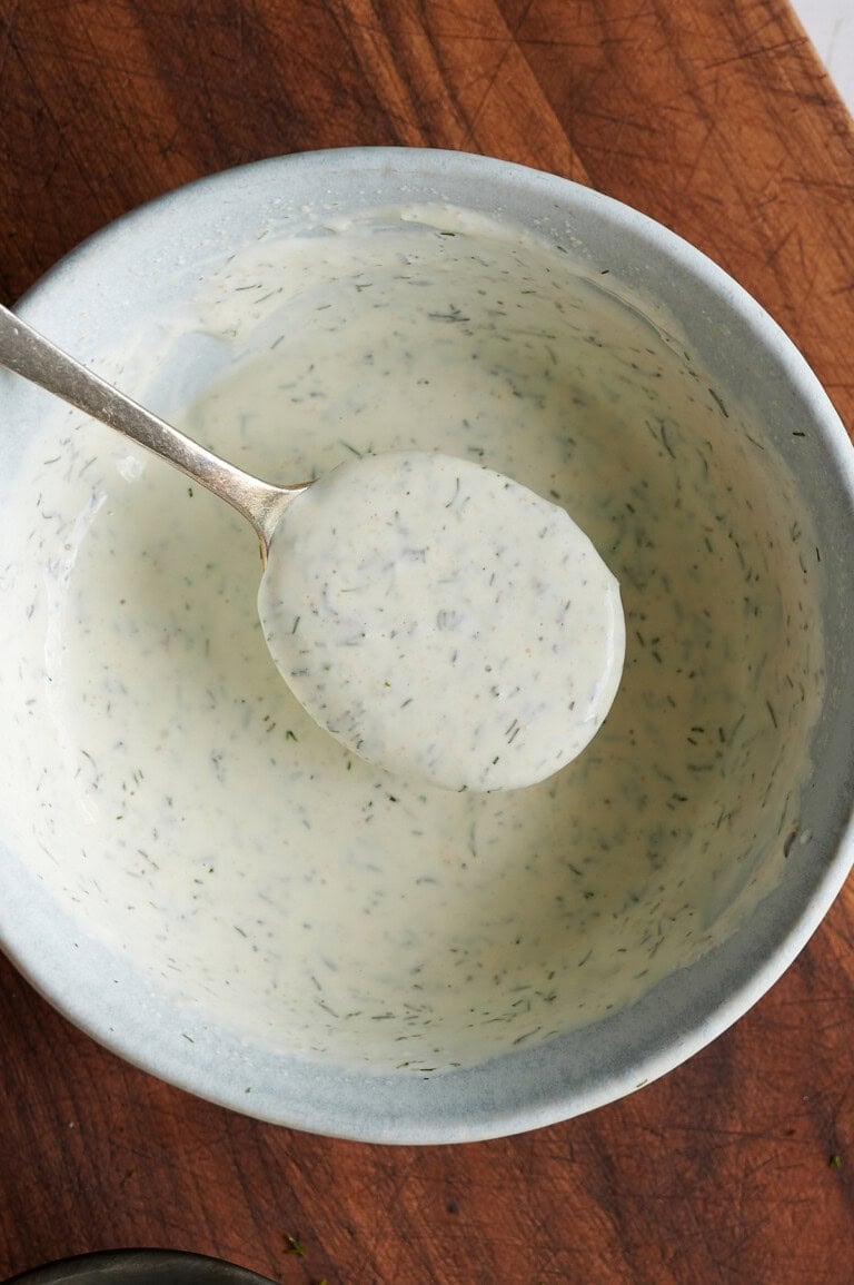 A spoon holding up creamy, herb-speckled ranch dressing over a light gray bowl on a wooden surface.