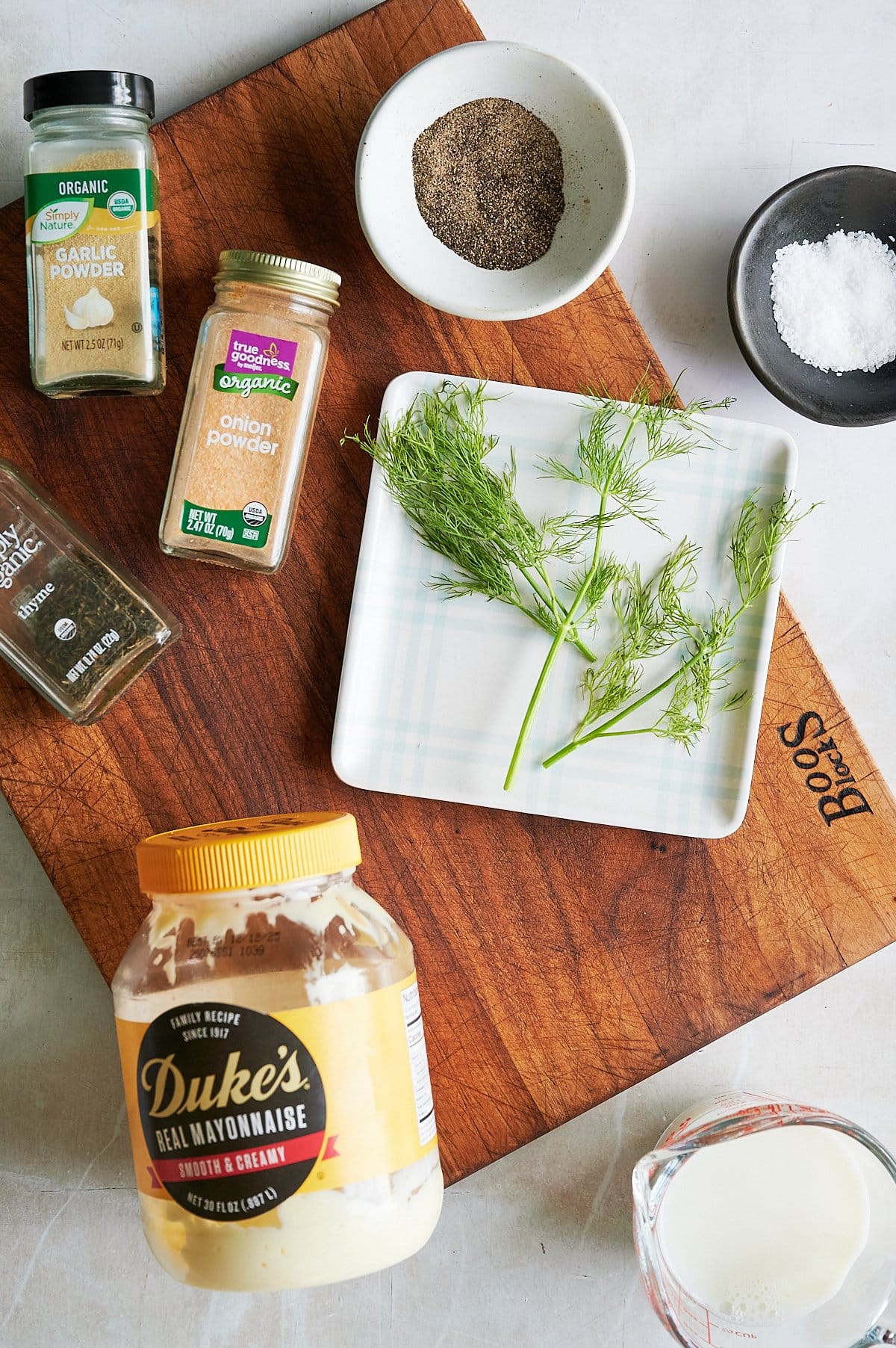 A wooden cutting board with fresh dill on a plate, a jar of mayonnaise, spices, a small bowl of salt, black pepper, and a small glass of milk for garlic ranch dressing.