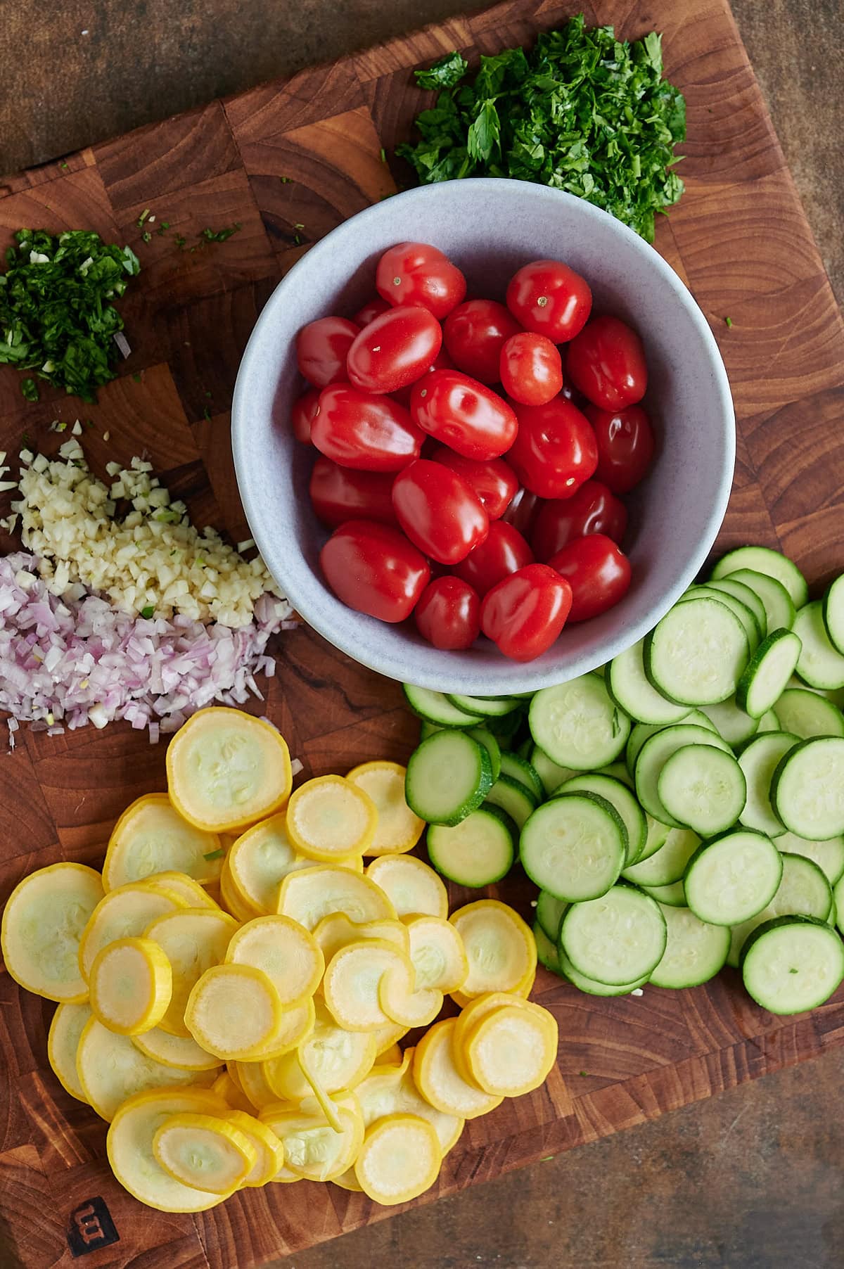 A bowl of grape tomatoes, sliced yellow squash, zucchini, chopped shallots, minced garlic, and herbs arranged on a wooden cutting board.