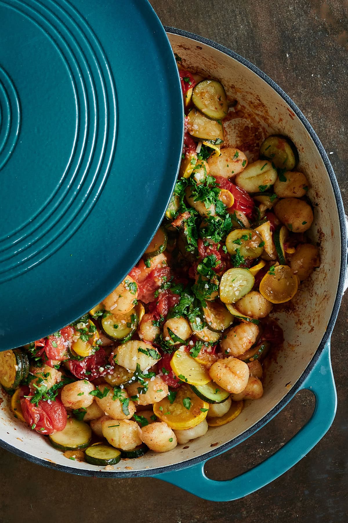 A blue Dutch oven partially covered, revealing a cooked dish of gnocchi, sliced zucchini, tomatoes, and herbs on a dark surface.