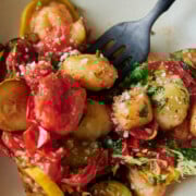A plate of gnocchi with tomato sauce, sliced zucchini, cherry tomatoes, grated cheese, and fresh herbs, with a black fork.