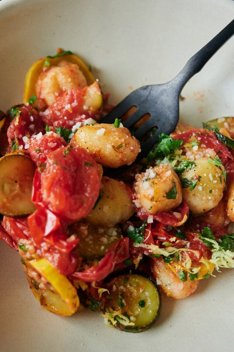 A plate of gnocchi with tomato sauce, sliced zucchini, cherry tomatoes, grated cheese, and fresh herbs, with a black fork.