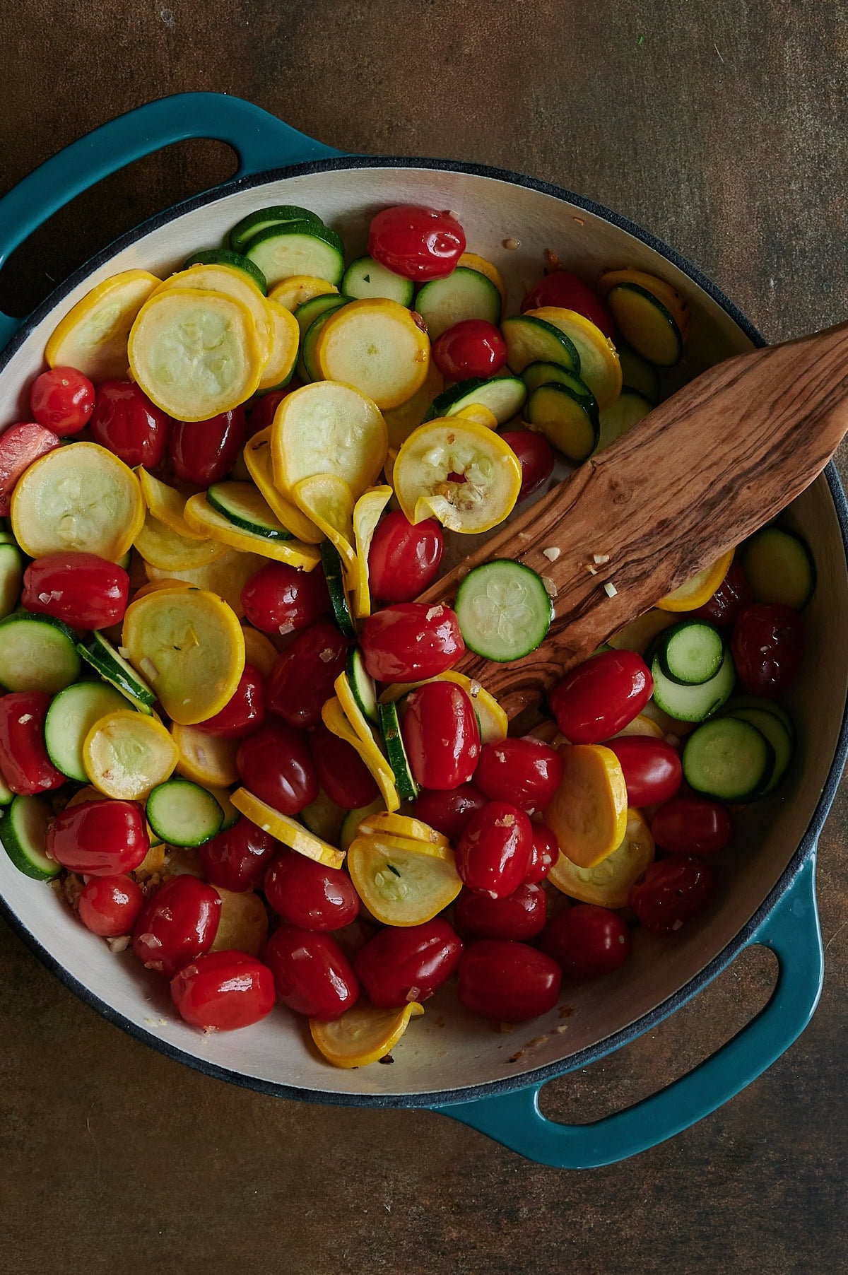 Sliced zucchini, yellow squash, and grape tomatoes being stirred with a wooden utensil in a blue-handled Dutch oven.