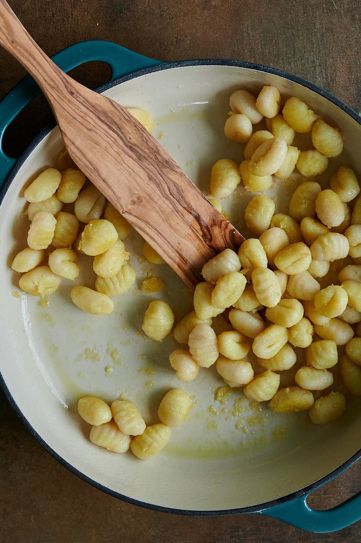A wooden spatula sautes gnocchi in a cream-colored pan with a teal rim, showing a light coating of oil.
