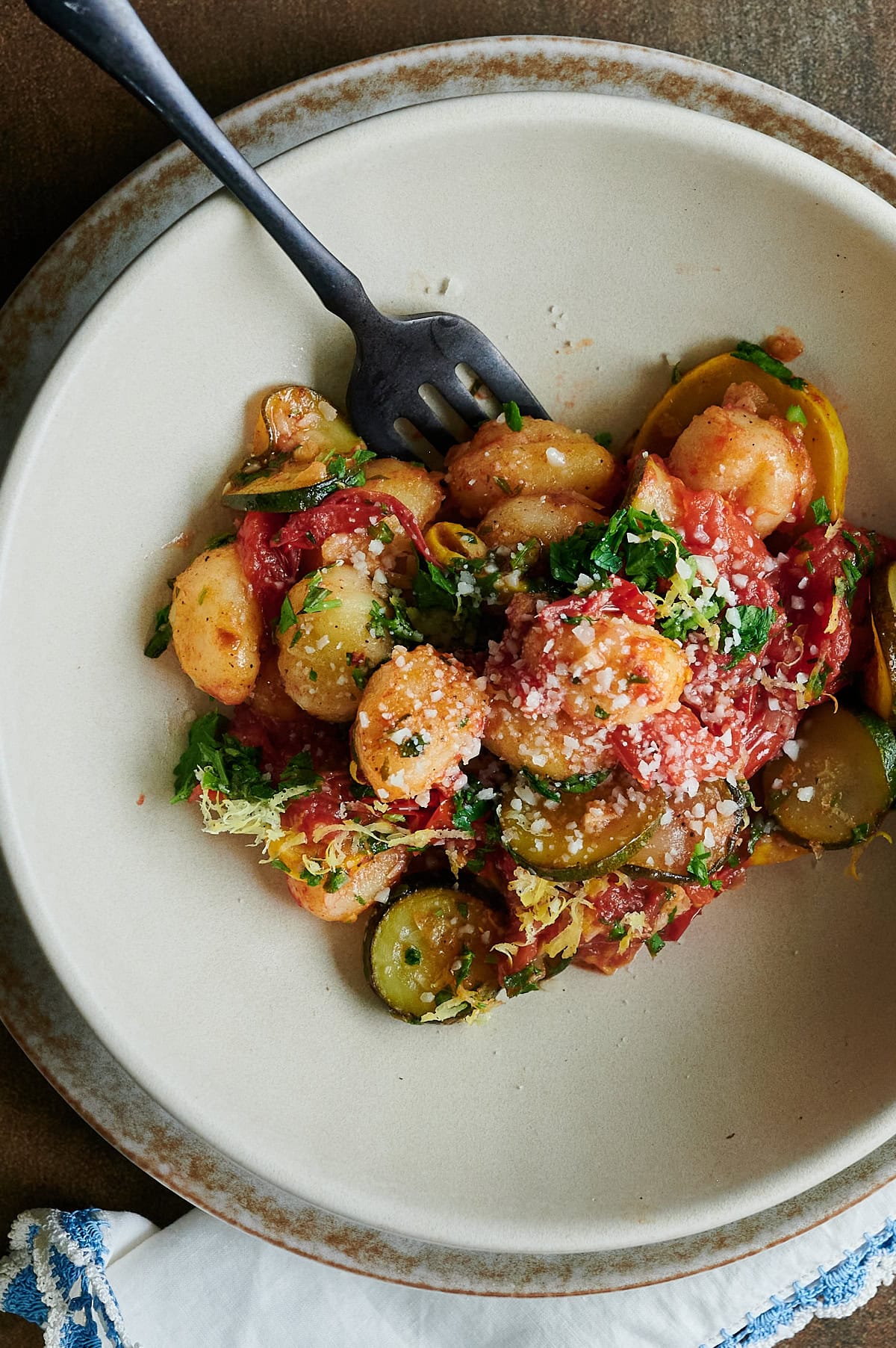 A plate of gnocchi with caramelized tomatoes, zucchini and yellow squash slices, grated cheese, and herbs, with a fork resting on the plate.
