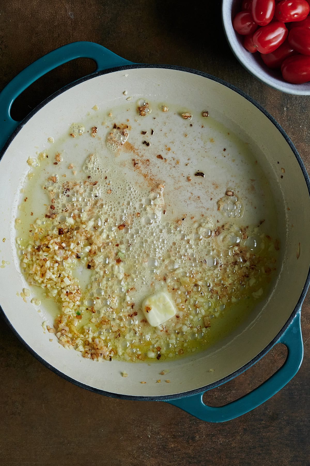 Chopped onions and garlic sautéing in melted butter in a large Dutch braiser; a bowl of grape tomatoes sits nearby.
