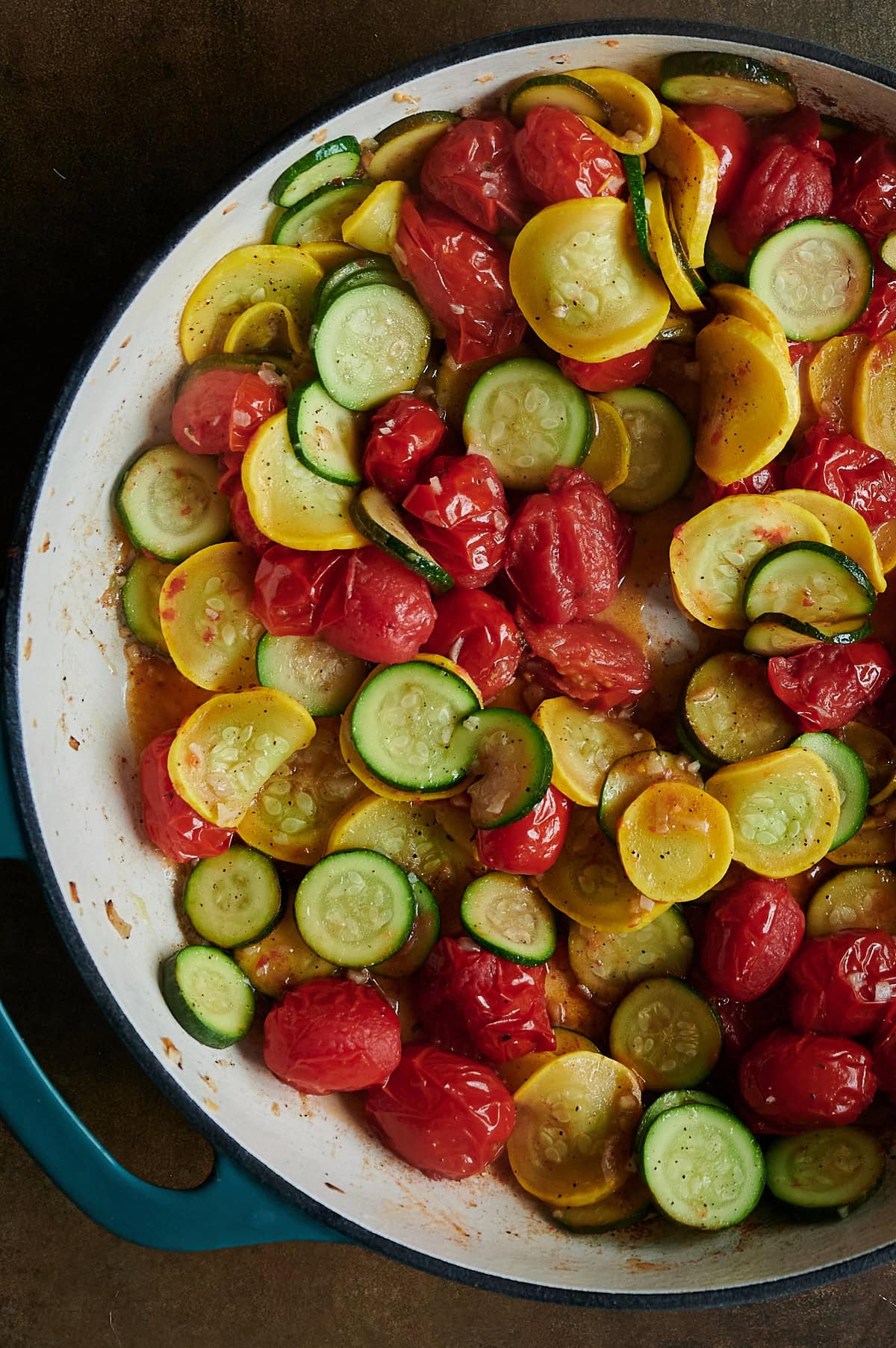 Sliced zucchini, yellow squash, and cherry tomatoes cooked in a large teal blue Dutch oven.