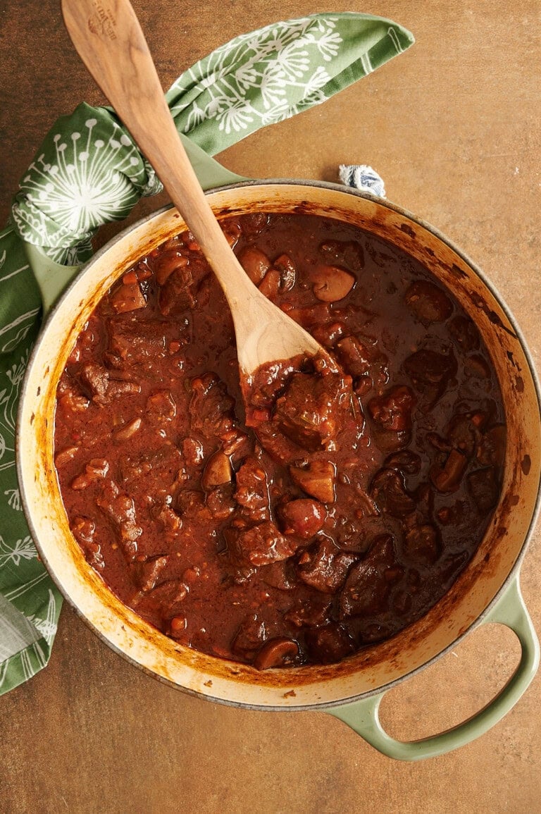 A pot of beef stew with chunks of meat and mushrooms in a rich brown sauce, being stirred with a wooden spoon on a brown surface.