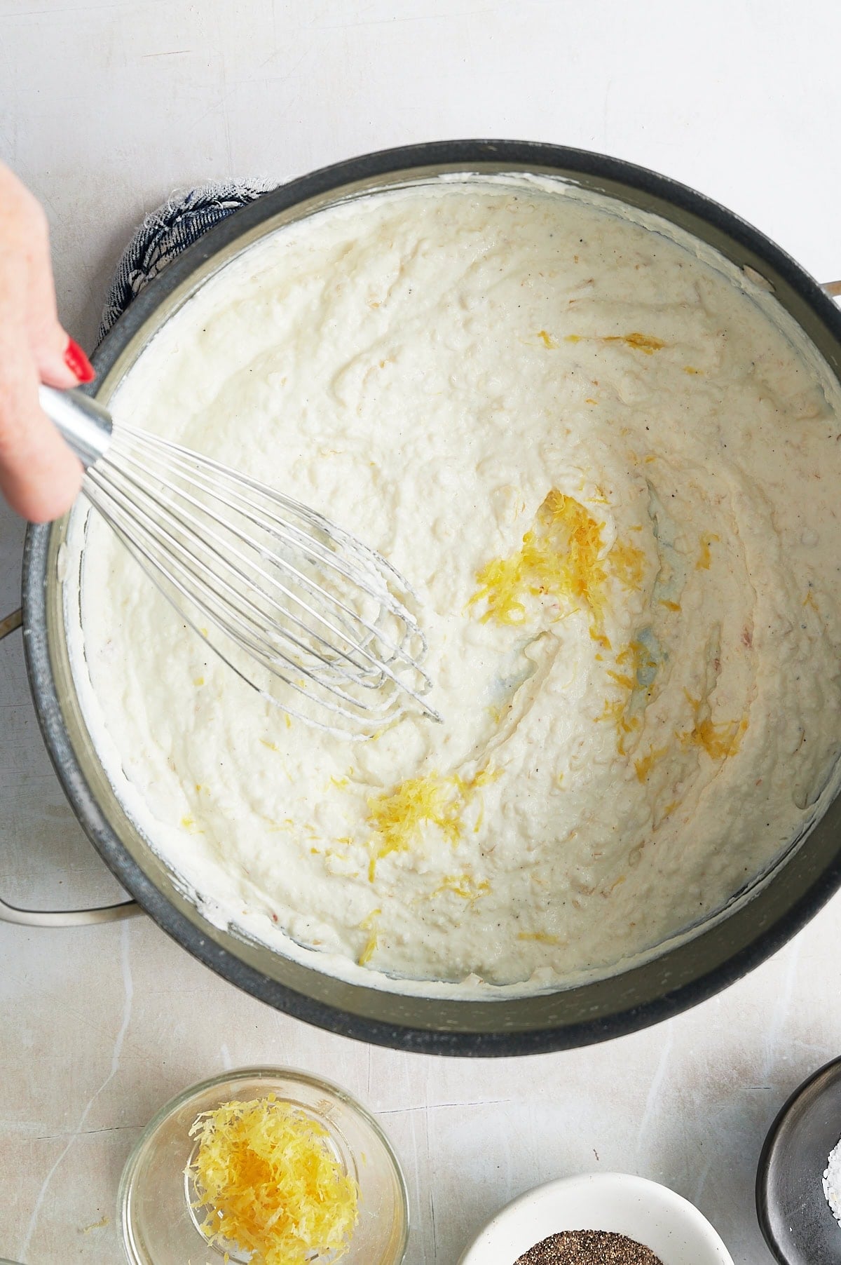 A hand whisking a creamy ricotta sauce with lemon zest in a pot, with small bowls of lemon zest and pepper nearby on a light surface.