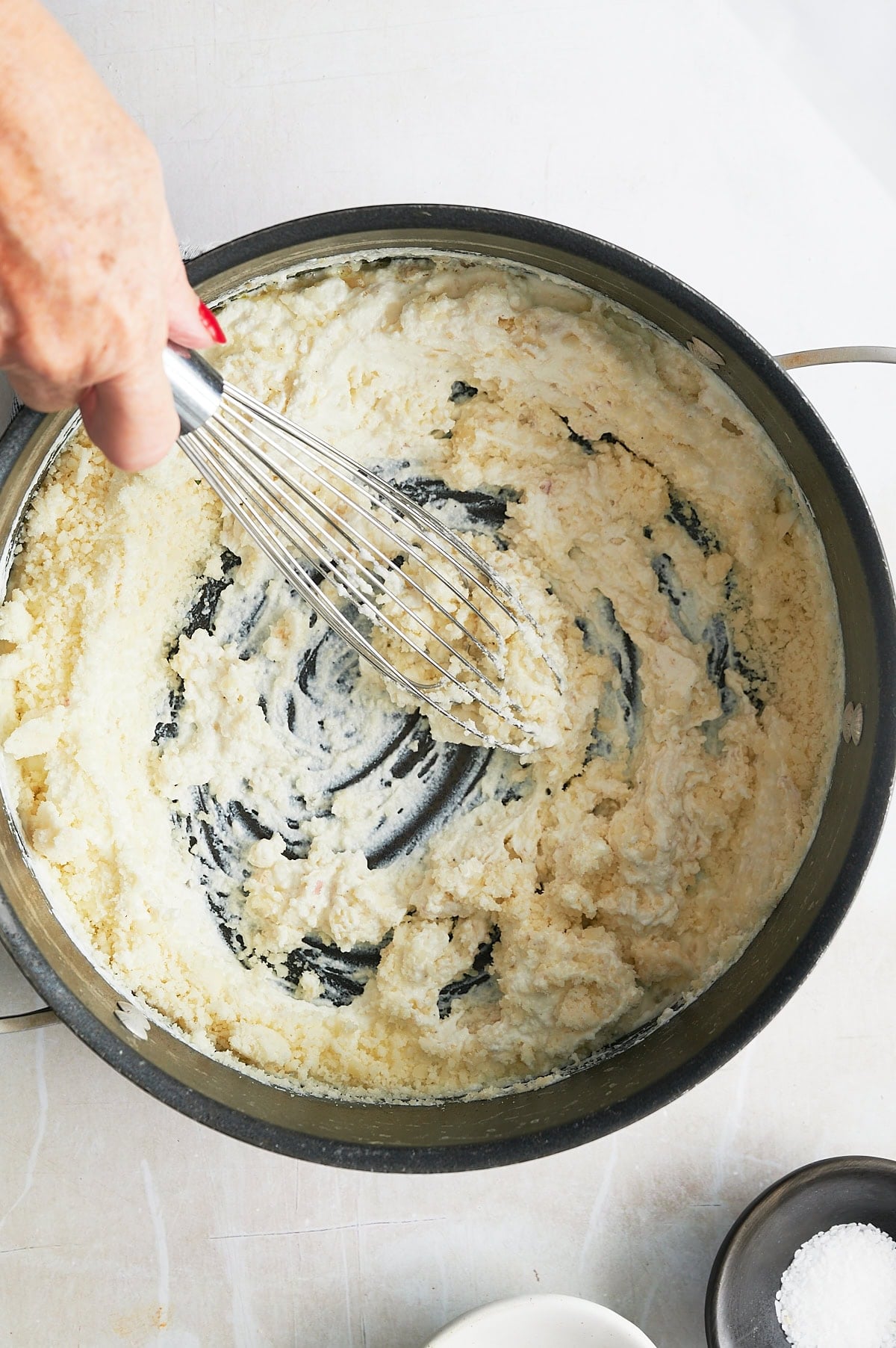 A hand using a whisk to stir a thick, creamy mixture in a black pan, with small bowls of salt and another ingredient nearby.