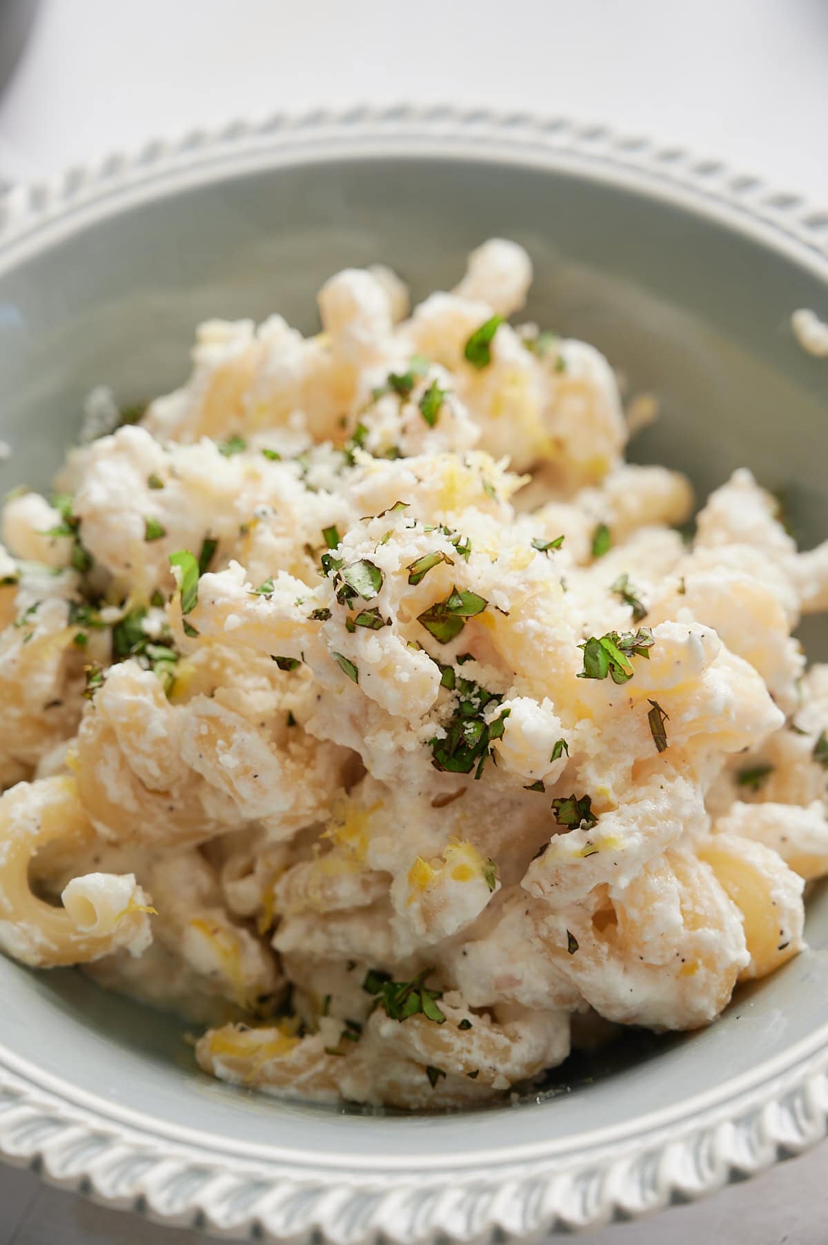 A bowl of creamy lemon ricotta pasta topped with chopped herbs and grated cheese, served in a light blue bowl with a decorative rim.