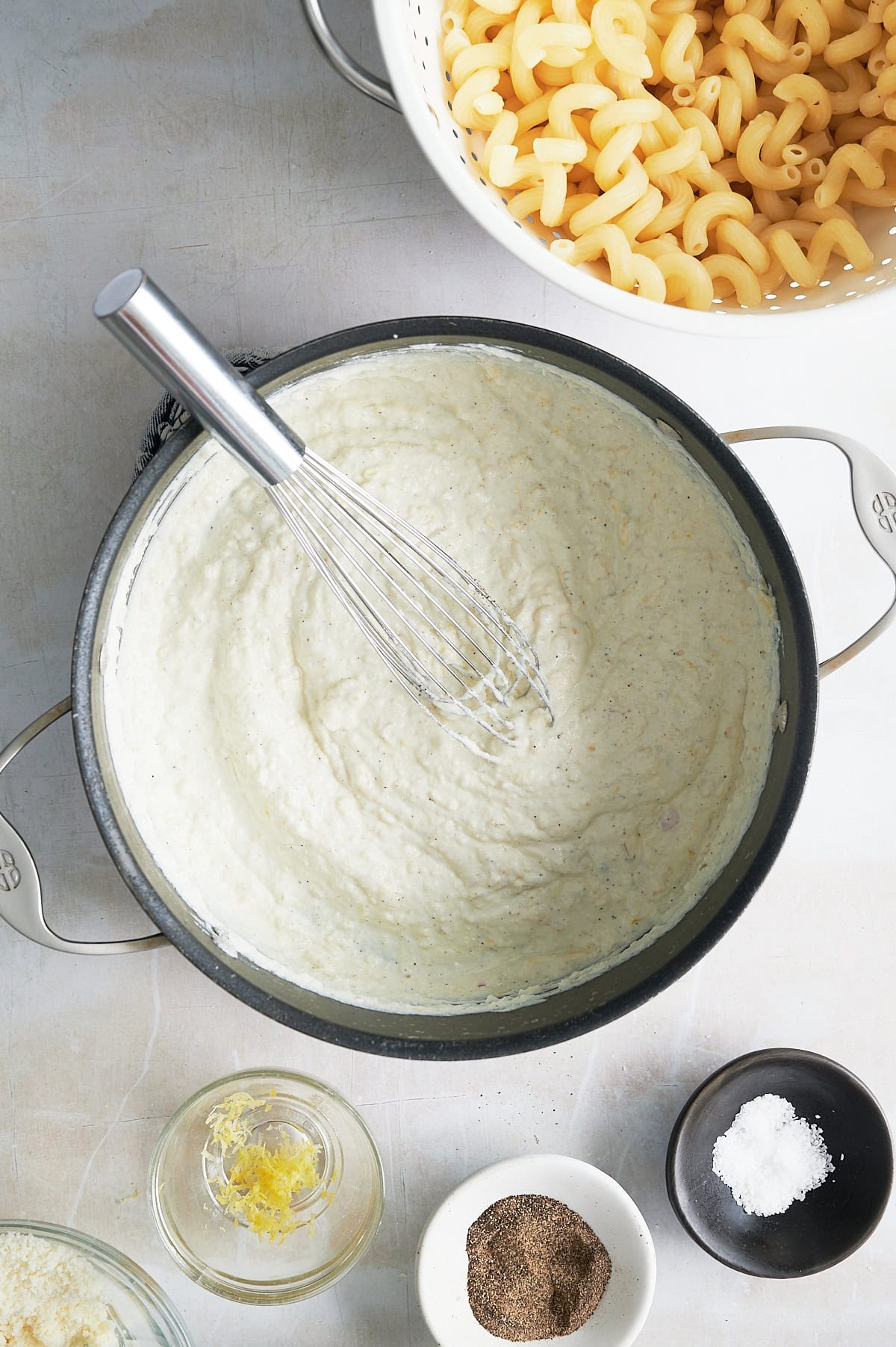 A pot of creamy ricotta cream sauce being whisked, with a colander of cooked pasta and small bowls of lemon zest, grated cheese, black pepper, and salt nearby.