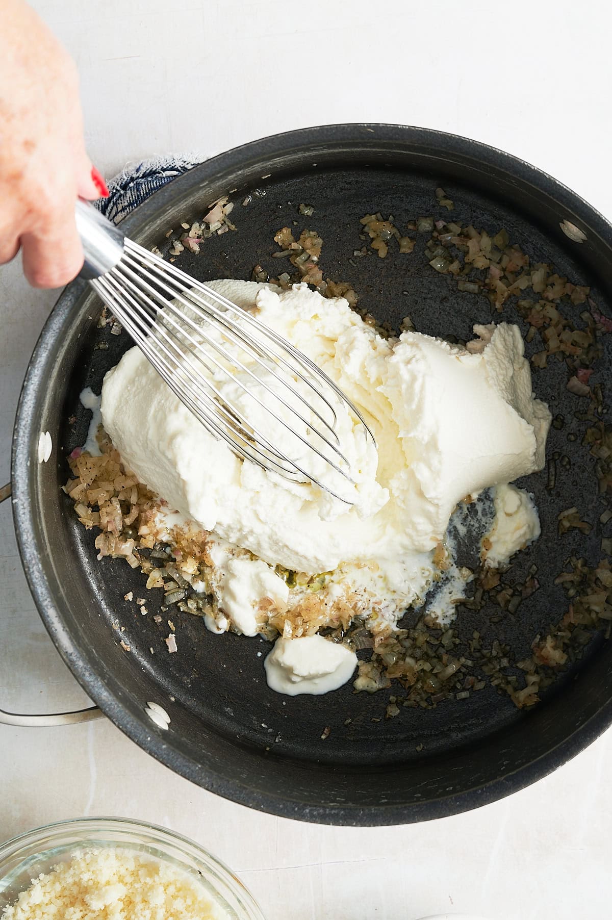 A hand uses a whisk to mix ricotta with sautéed onions and garlic in a black skillet.