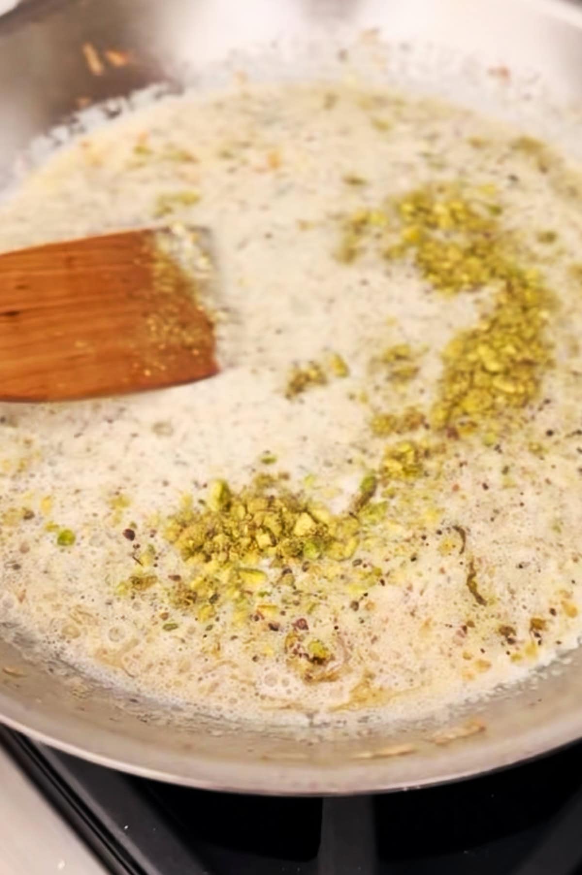 A close-up of a saucepan with a bubbling mixture of cream and chopped pistachios being stirred with a wooden spatula.
