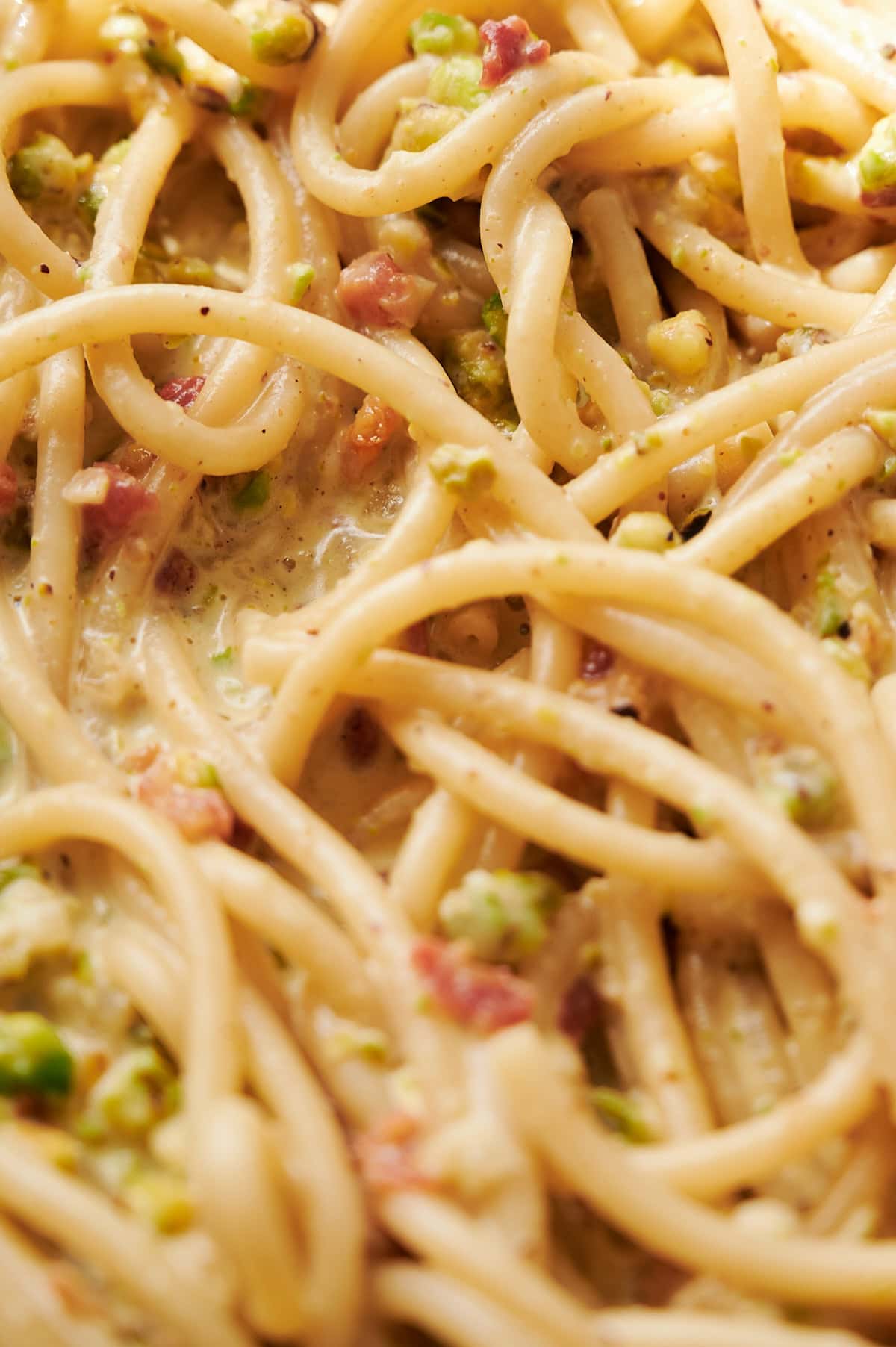 Close-up of cooked spaghetti mixed with a creamy sauce containing pistachios and small pieces of pancetta.