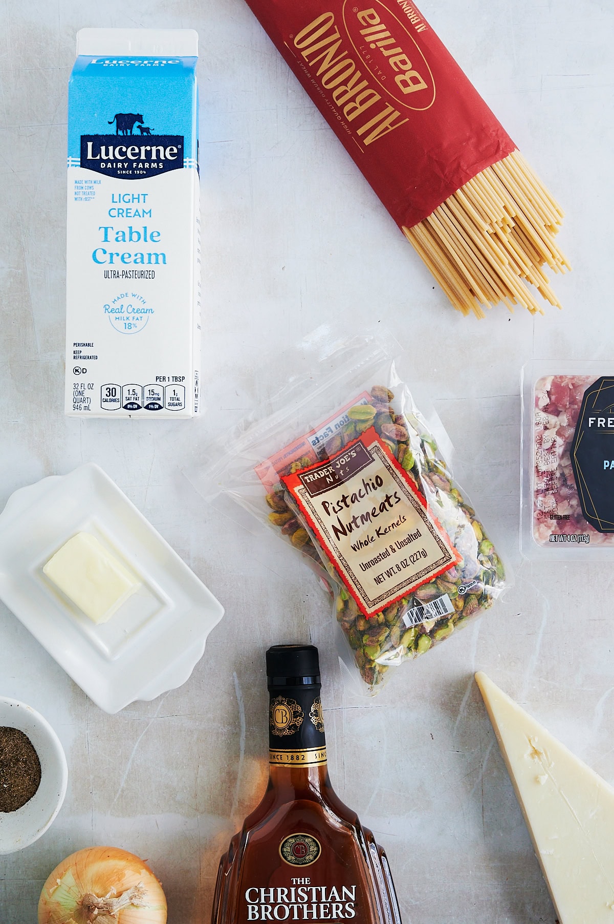 A carton of table cream, a package of bucatini, a bag of pistachios, a stick of butter, a wedge of cheese, diced pancetta, a bottle of brandy, a yellow onion, and a dish of pepper for pistachio sauce.