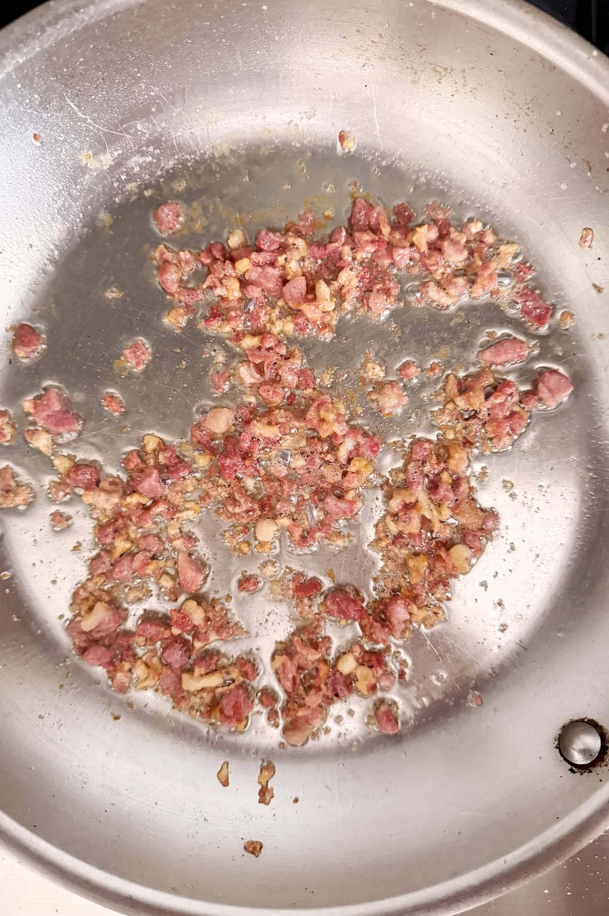 Ground Italian pancetta is browning in a stainless steel skillet on a stove, with some pieces starting to turn brown while others remain pink.