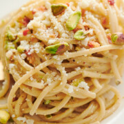 Close-up of a plate of spaghetti topped with grated cheese, crushed pistachios, and small pieces of pancetta.
