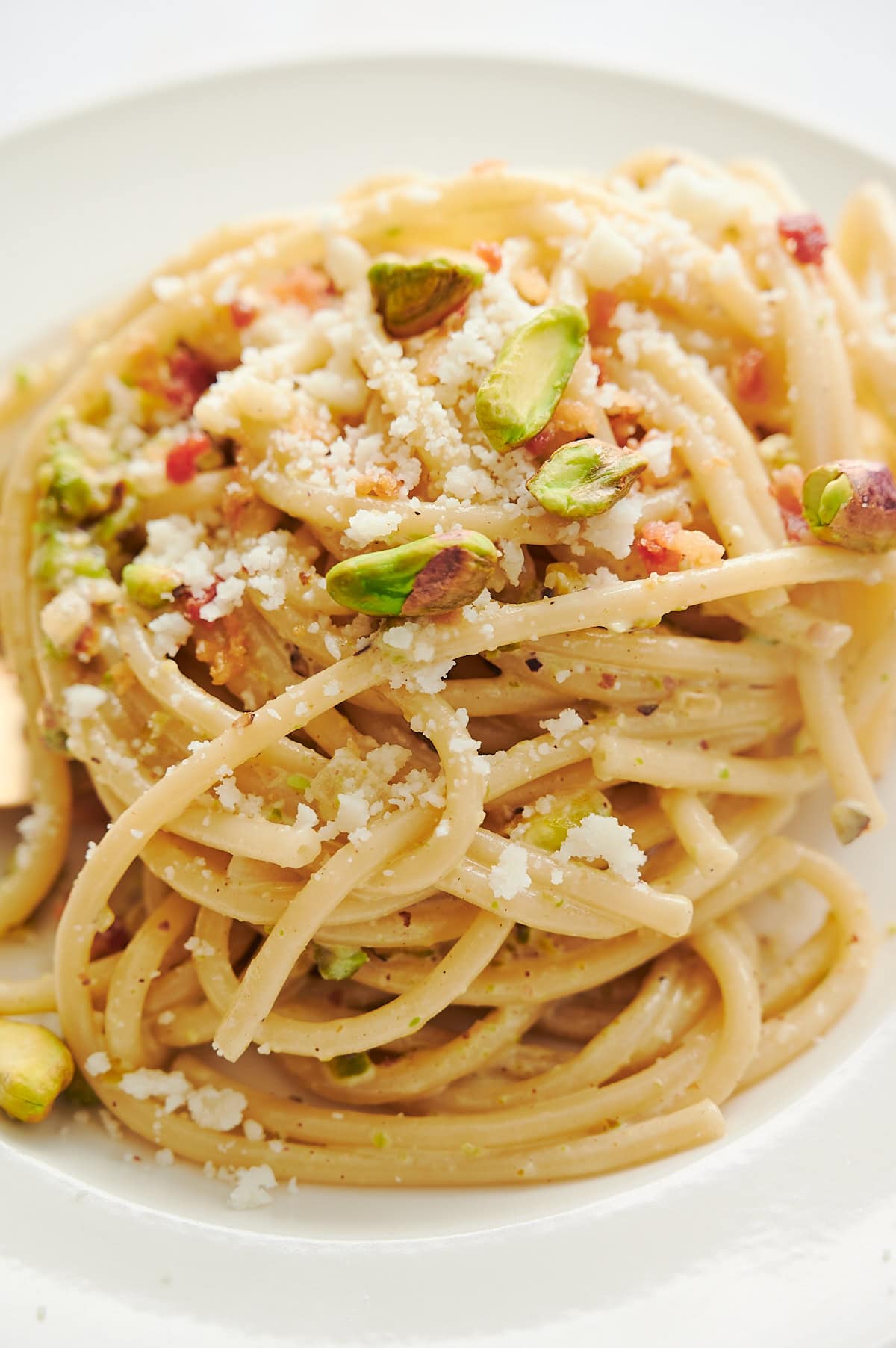 Close-up of a plate of spaghetti topped with grated cheese, crushed pistachios, and small pieces of pancetta.