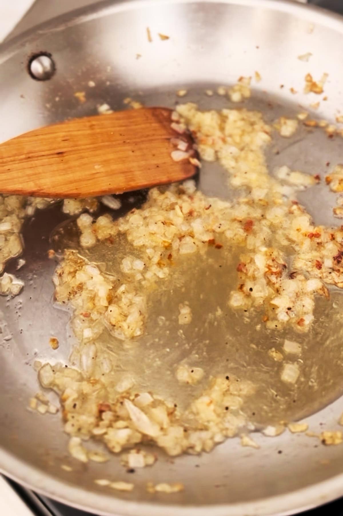 Chopped onions are sautéing in a stainless steel pan with brandy, being stirred with a wooden spatula.