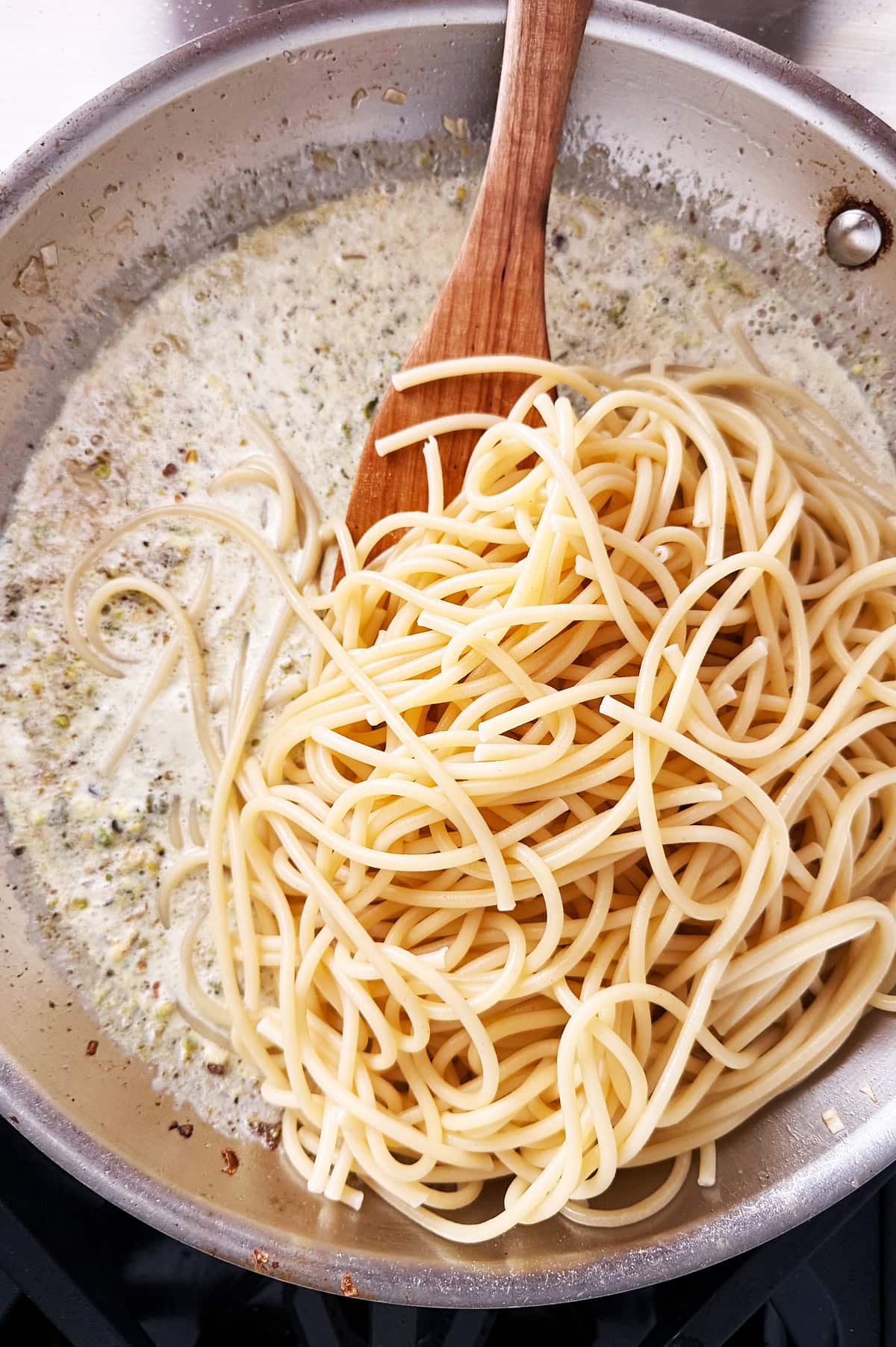Cooked bucatini being mixed into a creamy sauce in a stainless steel pan with a wooden spoon.