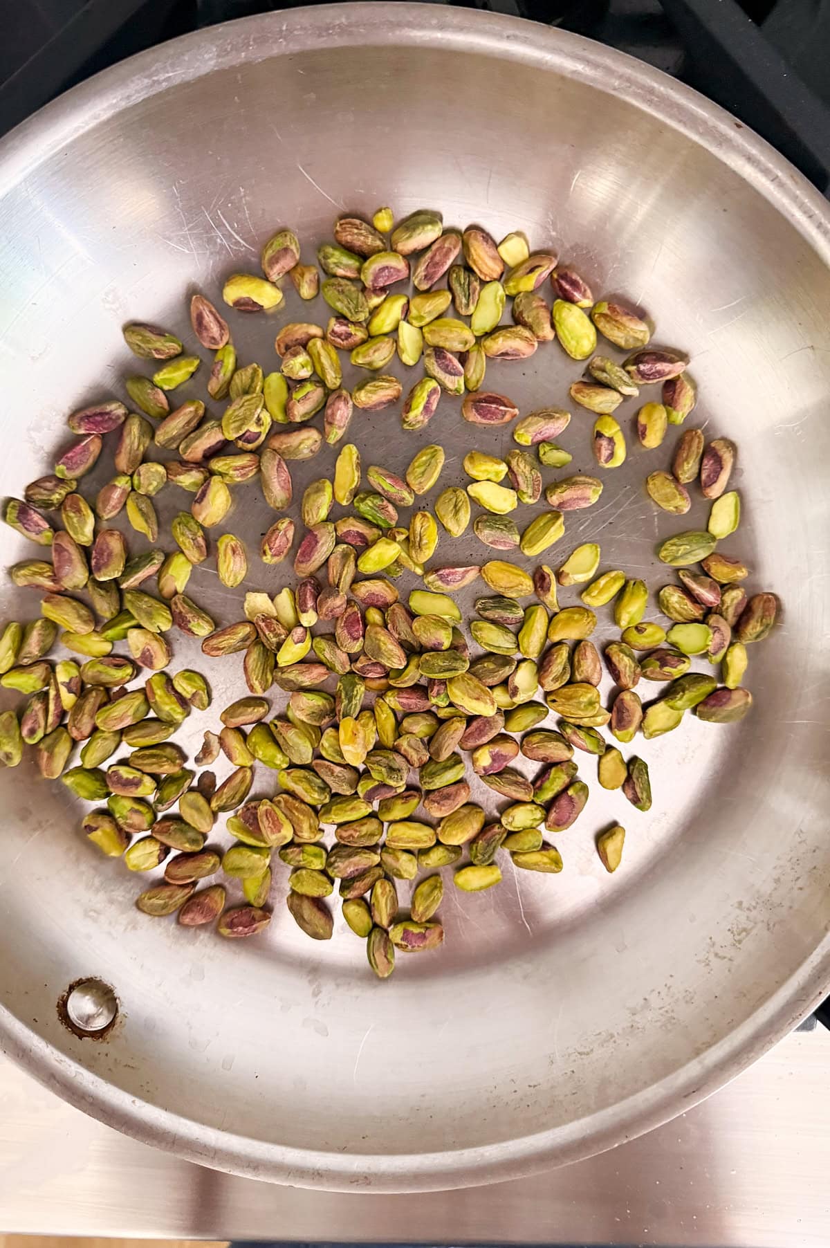 Shelled pistachios scattered in a single layer on a stainless steel pan, being toasted on a stovetop.