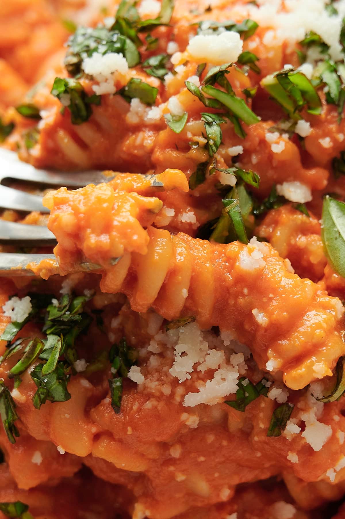 Close-up of rotini pasta with tomato sauce, garnished with chopped herbs and grated cheese, with a fork lifting a portion.
