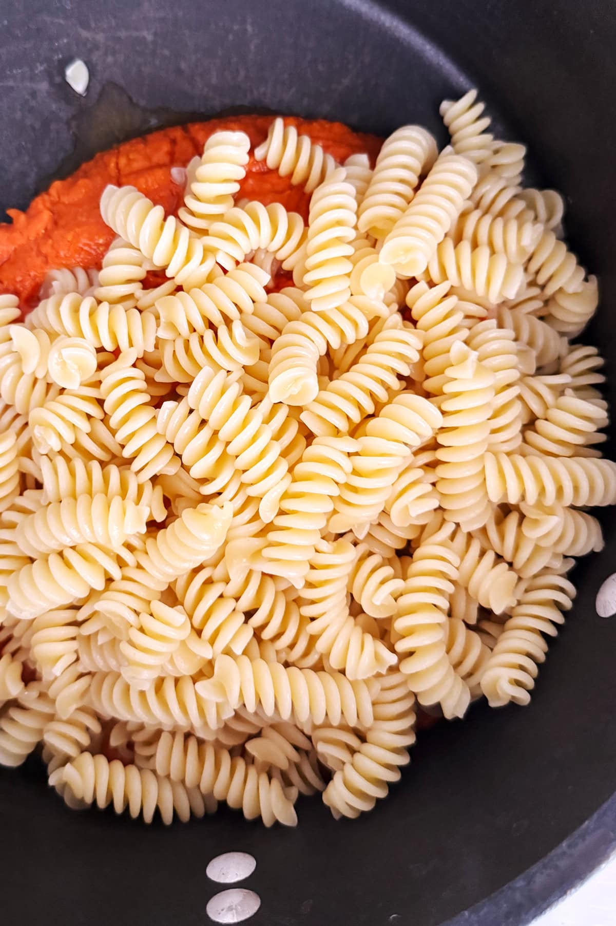 Cooked rotini pasta sits on top of cherry tomato sauce in a black pot, ready to be mixed together.