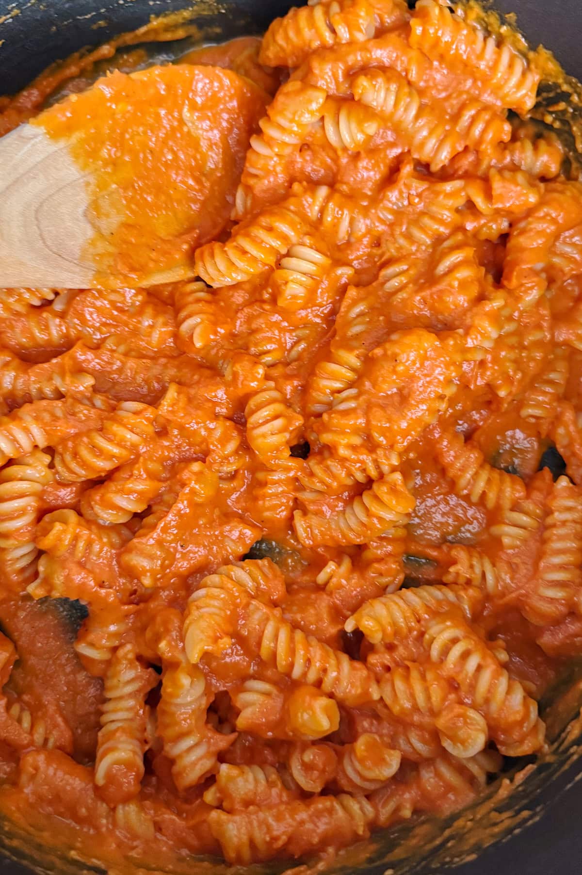 Cooked rotini pasta mixed with orange-red cherry tomato sauce, stirred with a wooden spoon in a pot.