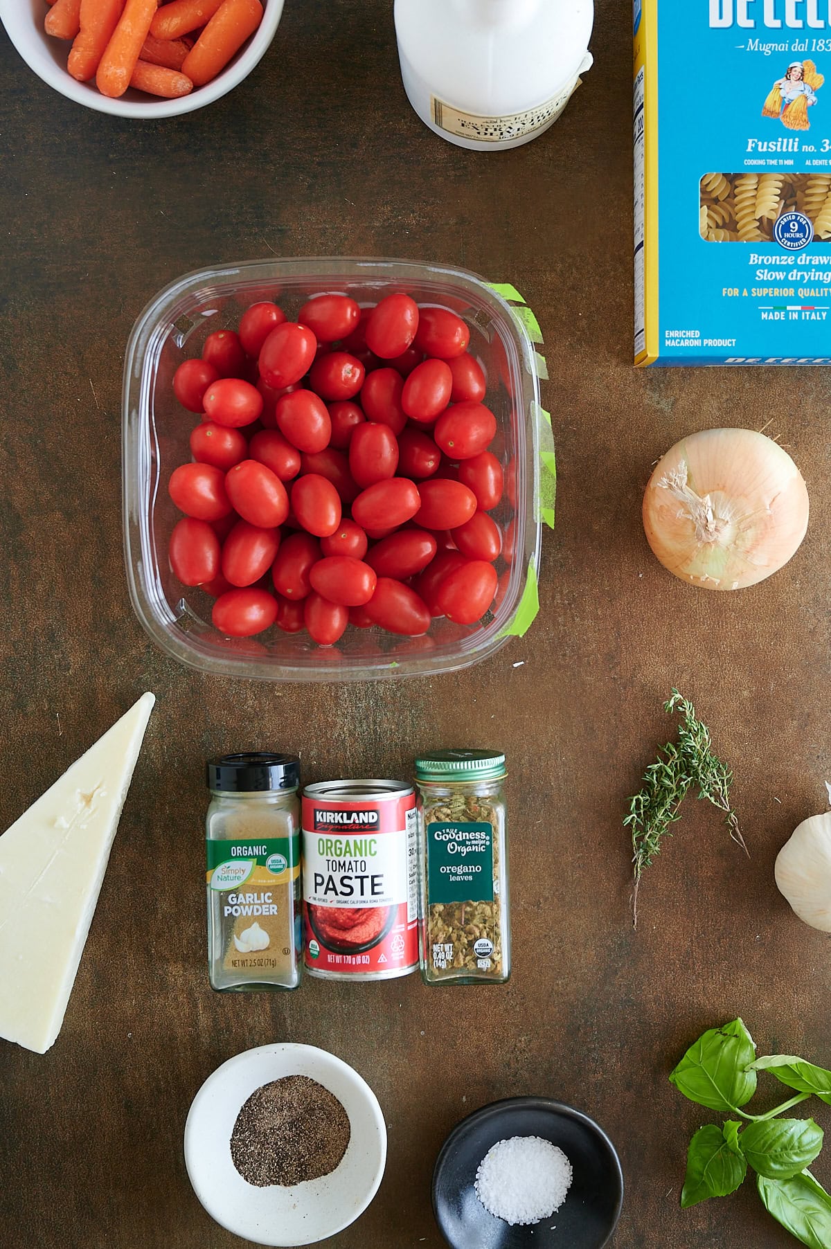 Assorted ingredients on a brown surface, including tomatoes, pasta, onion, herbs, seasonings, cheese, baby carrots, milk, and tomato paste.