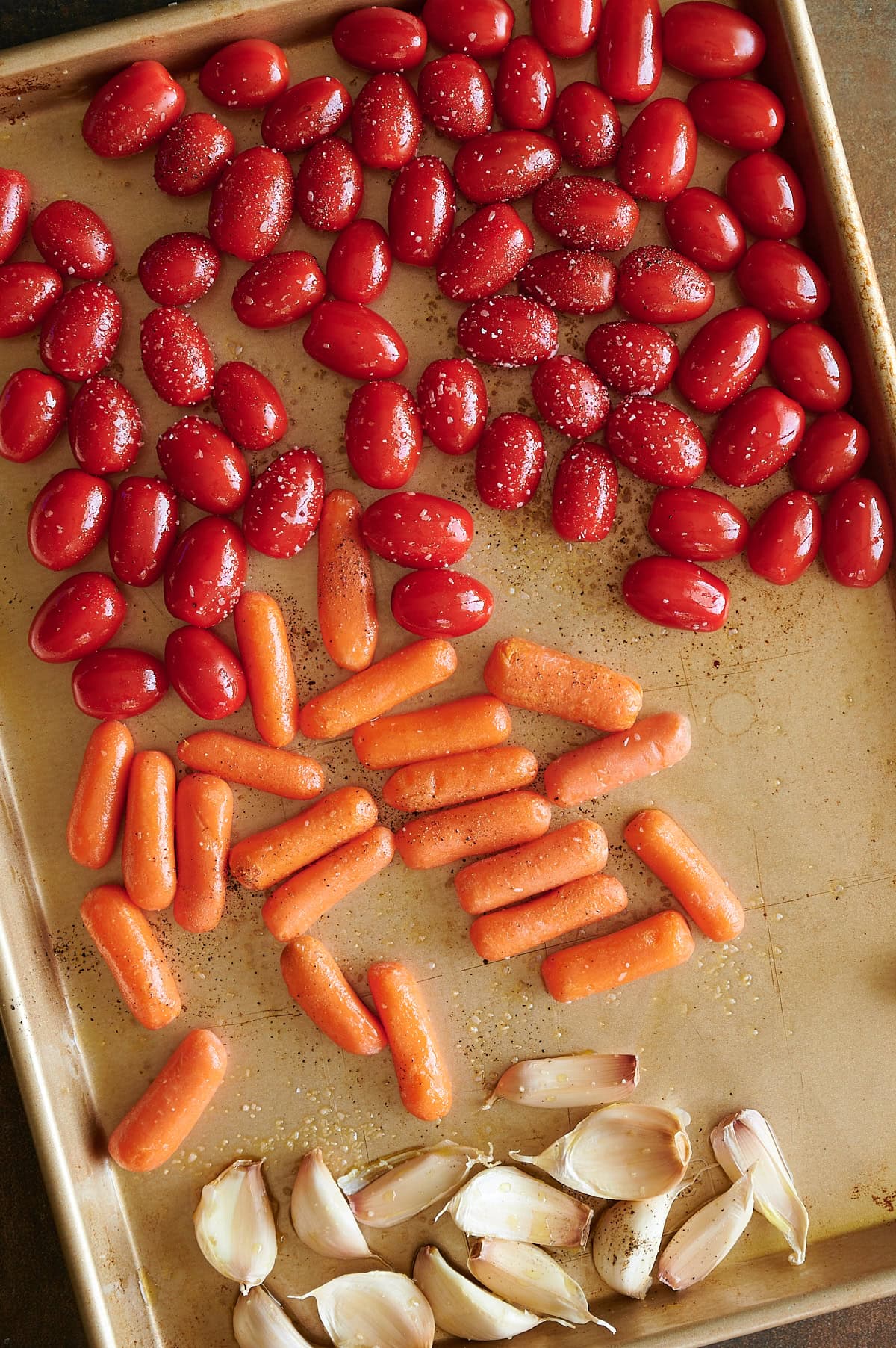 A baking sheet with cherry and grape tomatoes, baby carrots, and garlic cloves arranged in sections, all seasoned with salt and pepper.