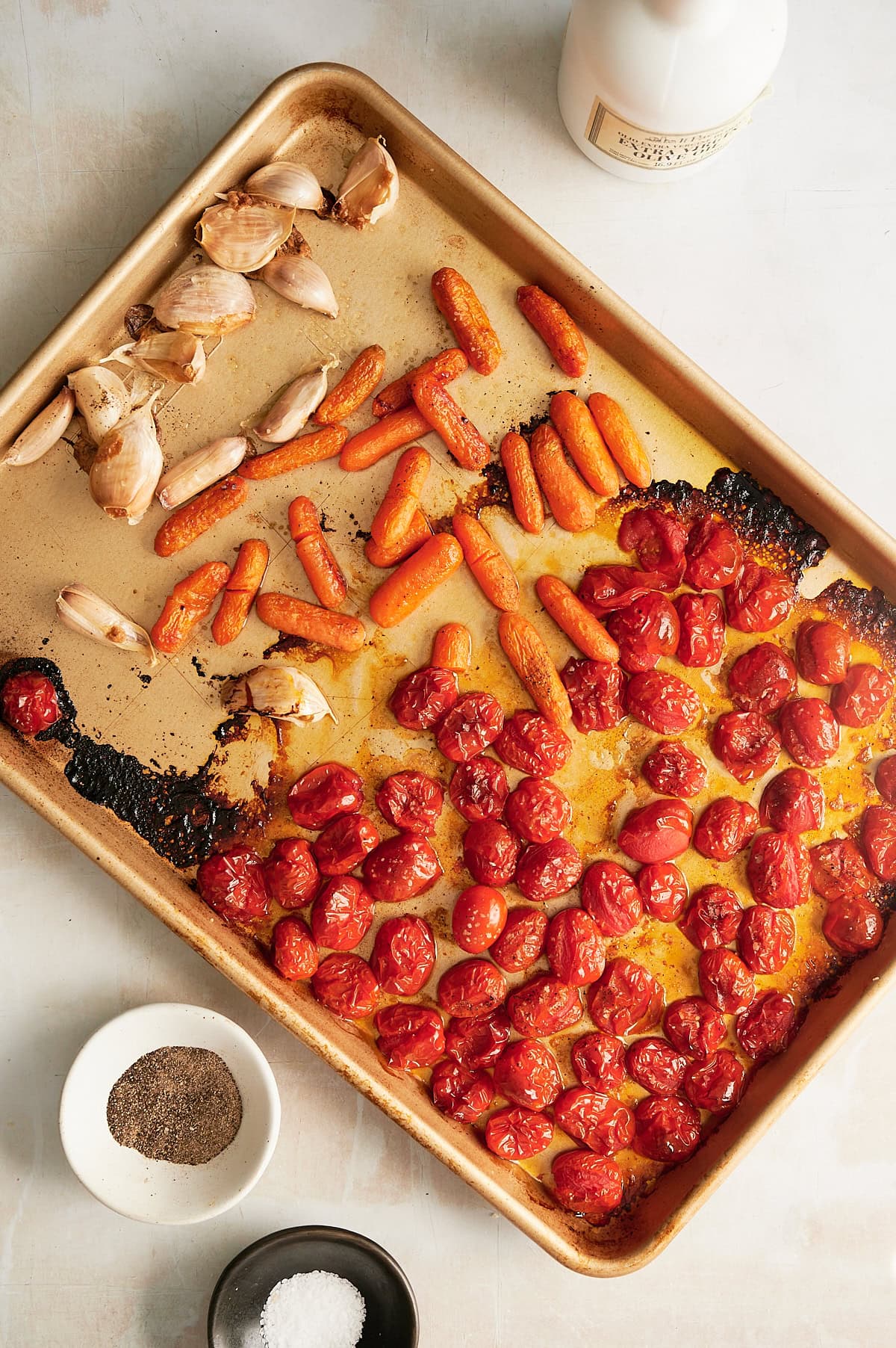 Roasted cherry tomatoes, baby carrots, garlic, and shallots on a baking sheet, with bowls of salt and pepper and a bottle nearby.