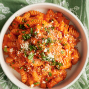 A bowl of ridged pasta in roasted tomato sauce, topped with grated cheese and chopped herbs, sits on a green patterned cloth with a spoon nearby.