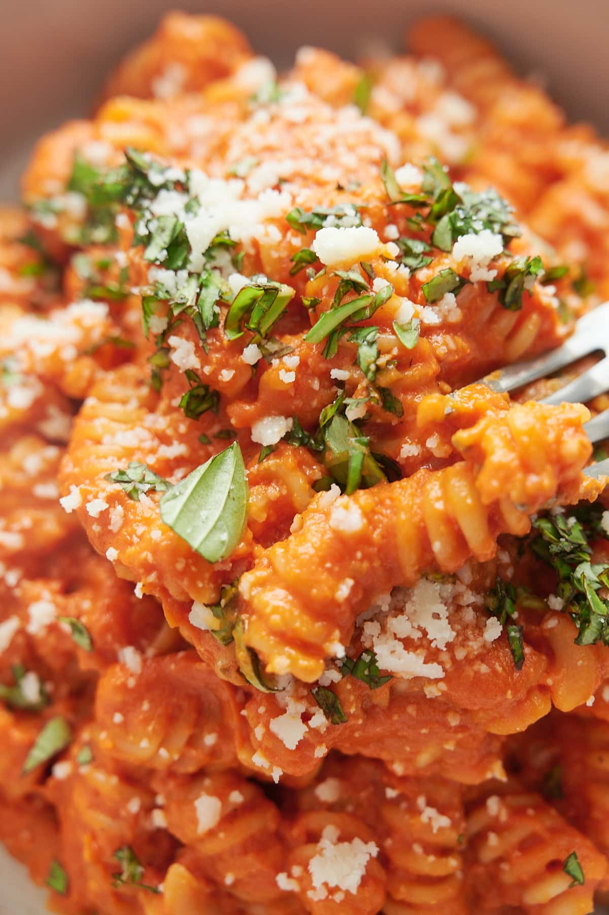 A close-up of rotini pasta covered in roasted cherry tomato sauce, topped with grated cheese and chopped fresh herbs, with a fork lifting a portion.