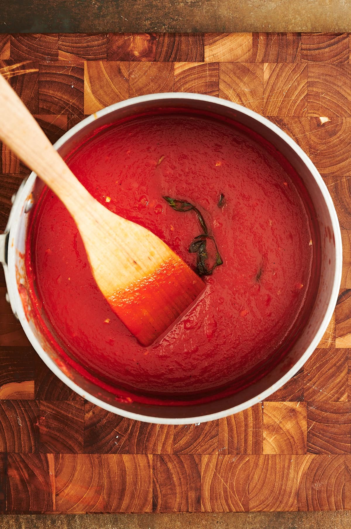 A saucepan of red tomato sauce with a wooden spoon rests on a wooden cutting board; basil leaves are visible in the sauce.