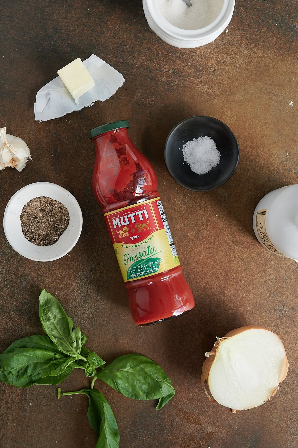 A bottle of Mutti tomato passata surrounded by butter, salt, pepper, sugar, garlic, basil leaves, and half an onion on a brown surface.
