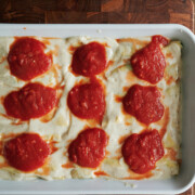 A baking dish of cannelloni topped with dollops of passata tomato sauce, set on a wooden cutting board with a pot of sauce nearby.