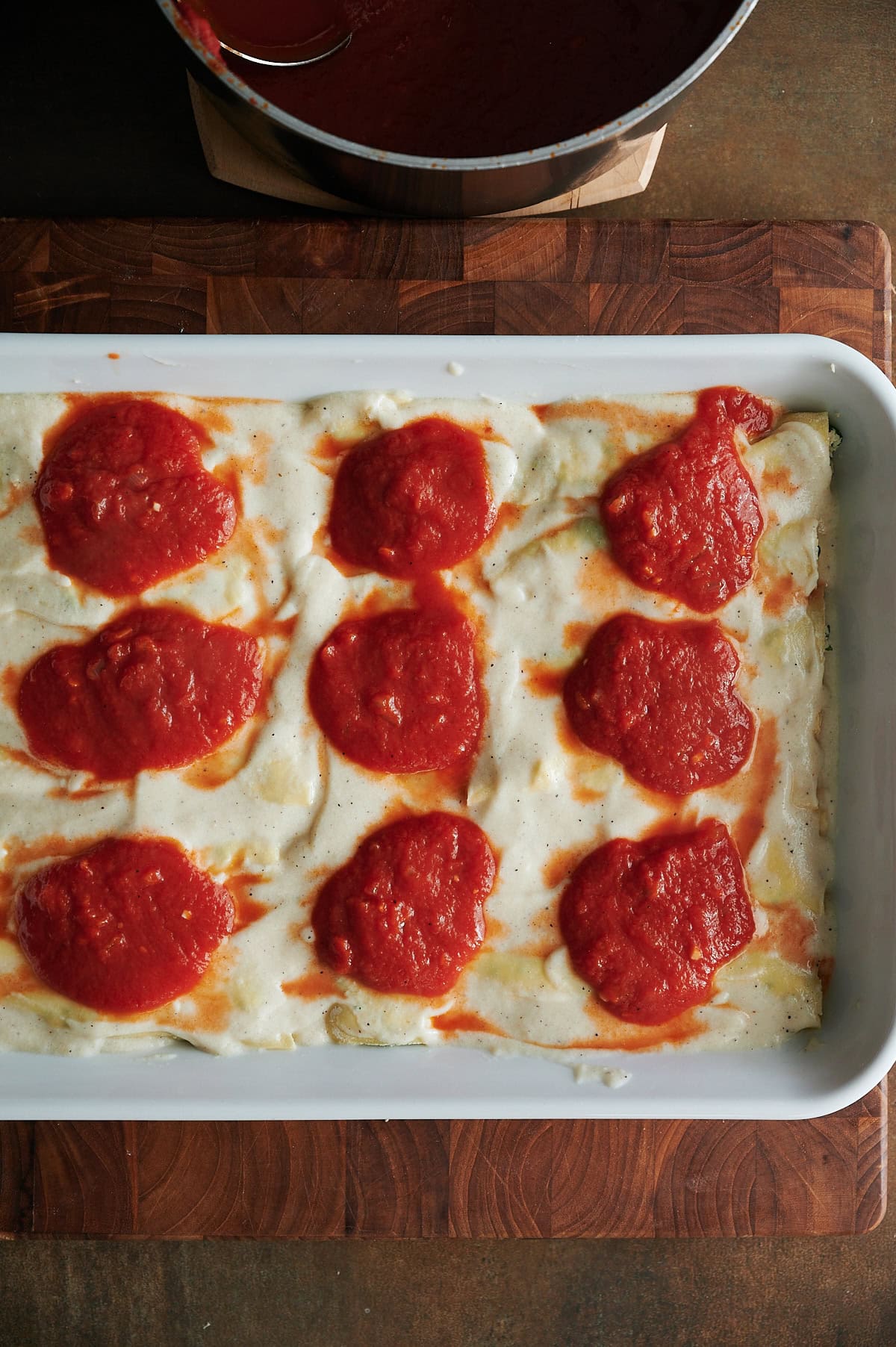 A baking dish of cannelloni topped with dollops of passata tomato sauce, set on a wooden cutting board with a pot of sauce nearby.