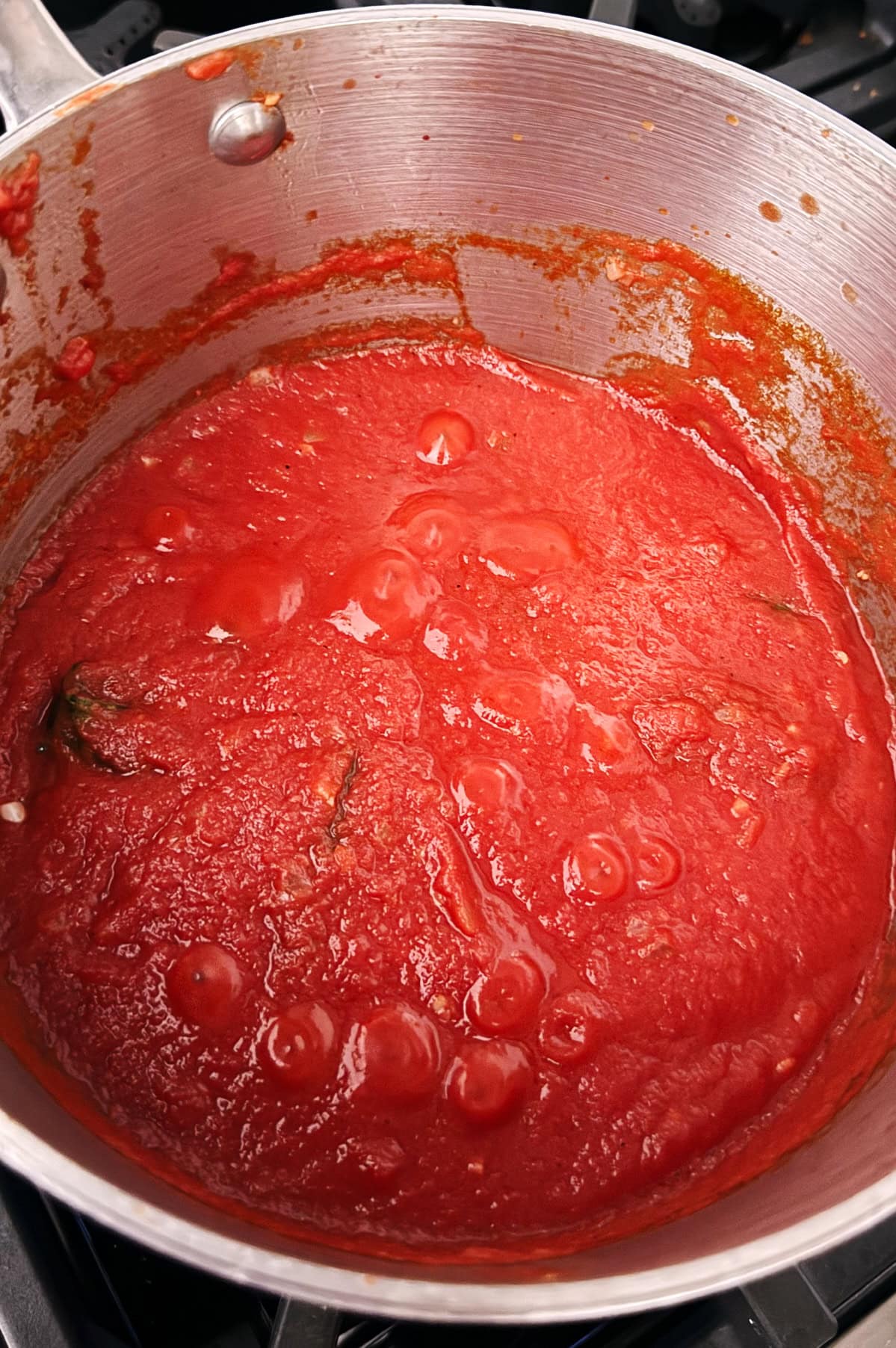 A stainless steel pot containing simmering passata tomato sauce with visible herbs on a stovetop.