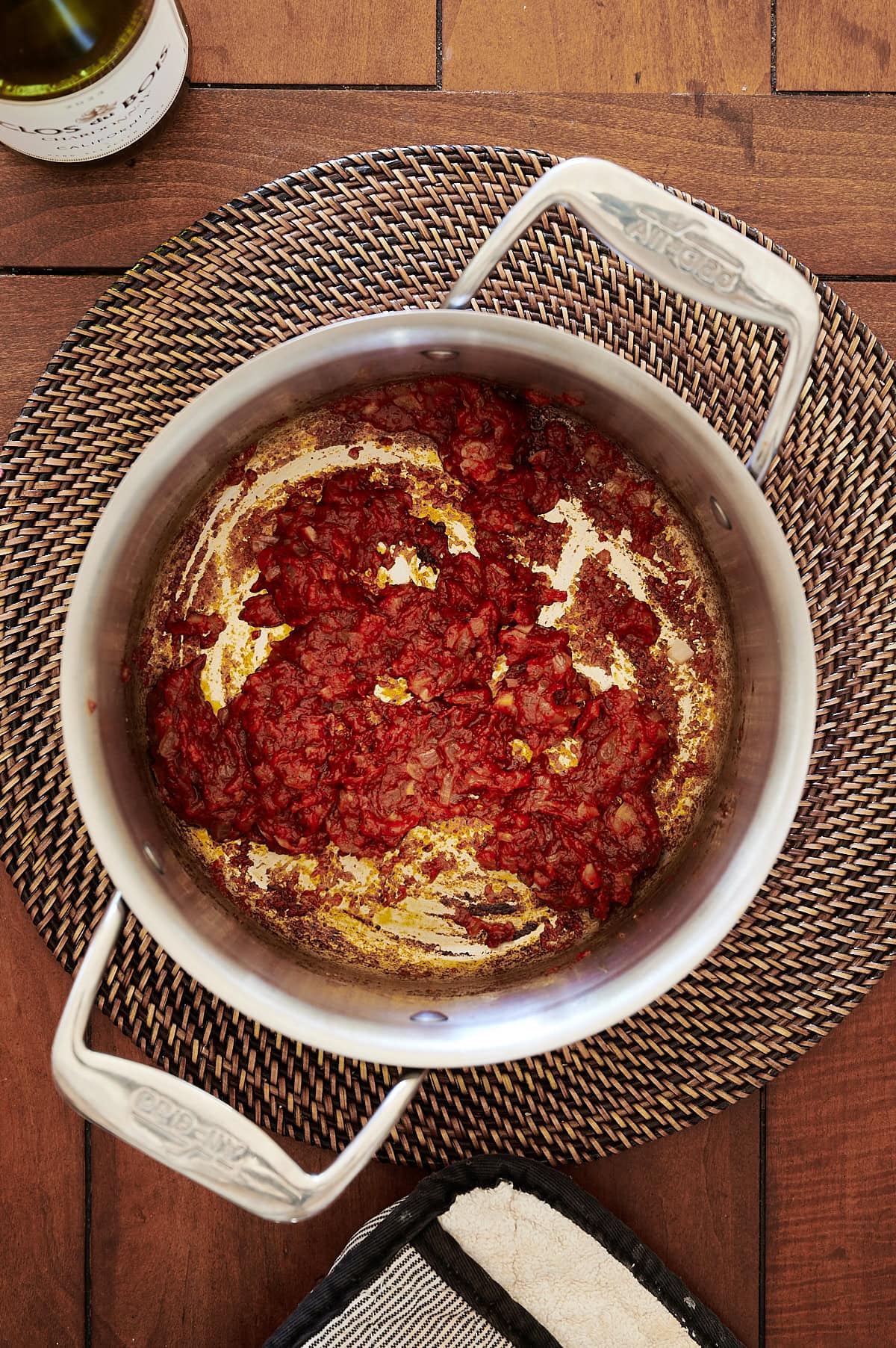 A stainless steel pot containing sautéed tomato paste sits on a woven placemat on a wooden table, with a bottle and a tray partially visible nearby.