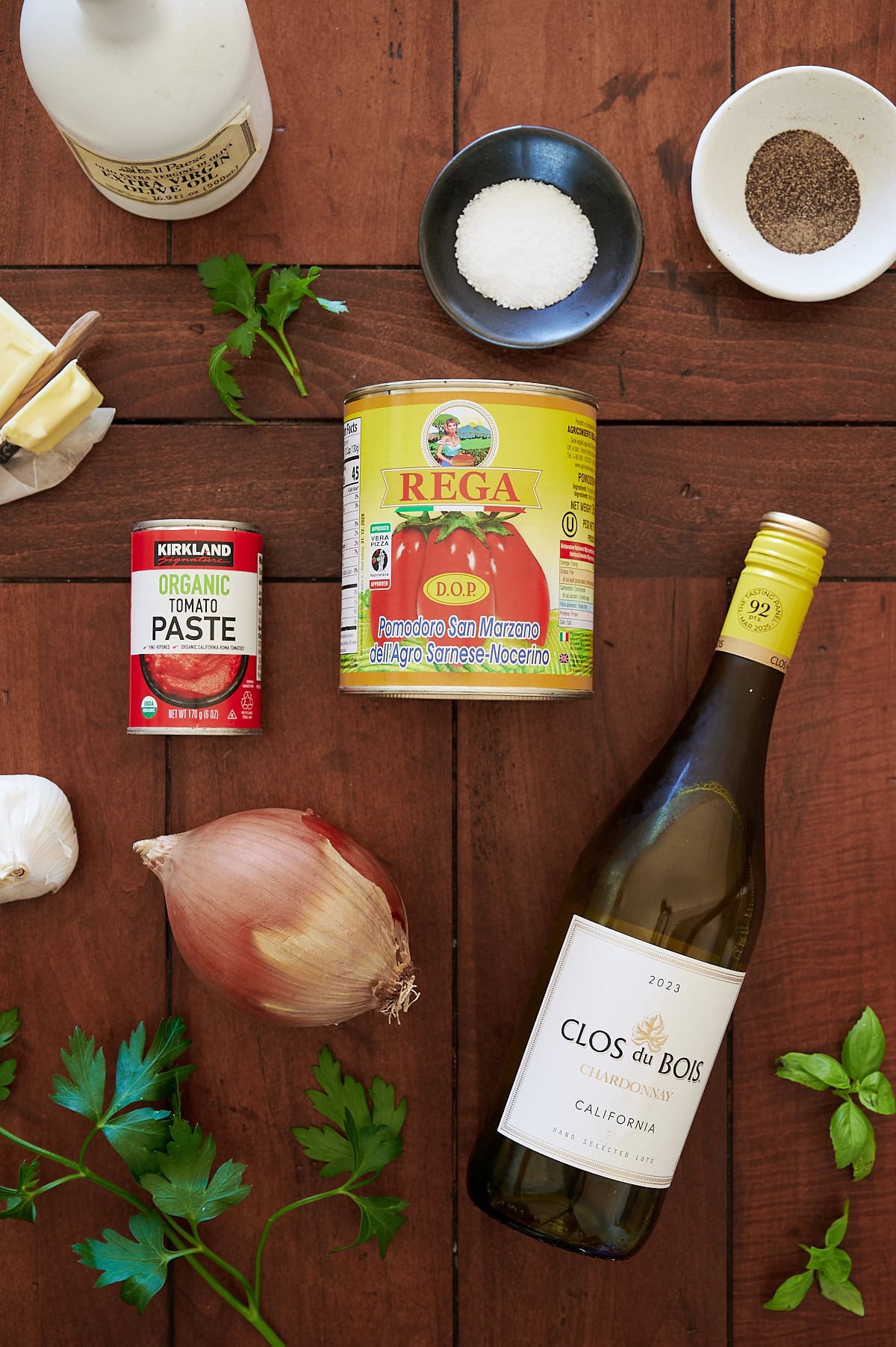 Ingredients for a recipe arranged on a wooden surface, including canned tomatoes, tomato paste, white wine, shallot, parsley, butter, salt, and pepper.