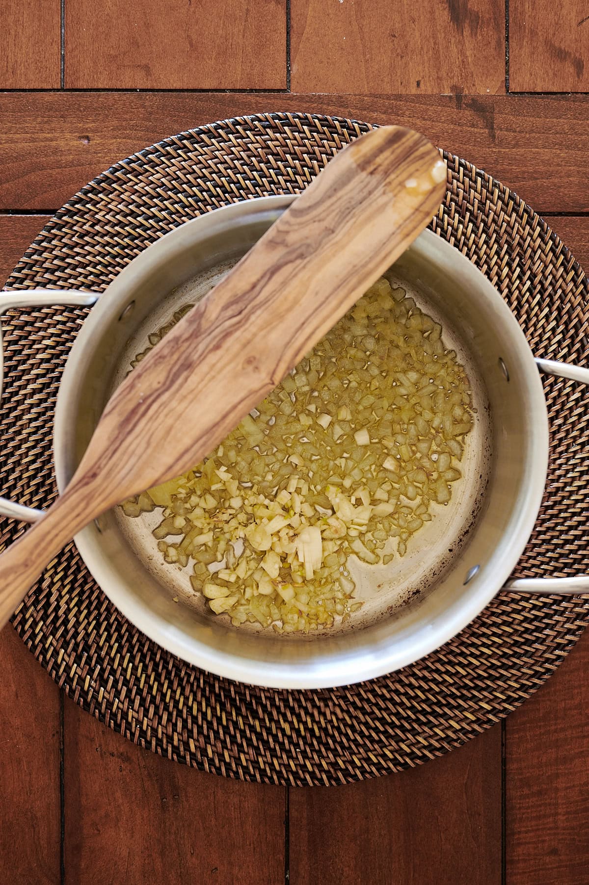 A wooden spatula rests on a pot with chopped onions and garlic being sautéed, placed on a woven mat atop a wooden table.