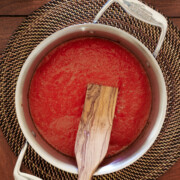 A saucepan filled with tomato sauce sits on a woven placemat, with a wooden spatula resting inside. A wine bottle is partially visible in the top left corner.