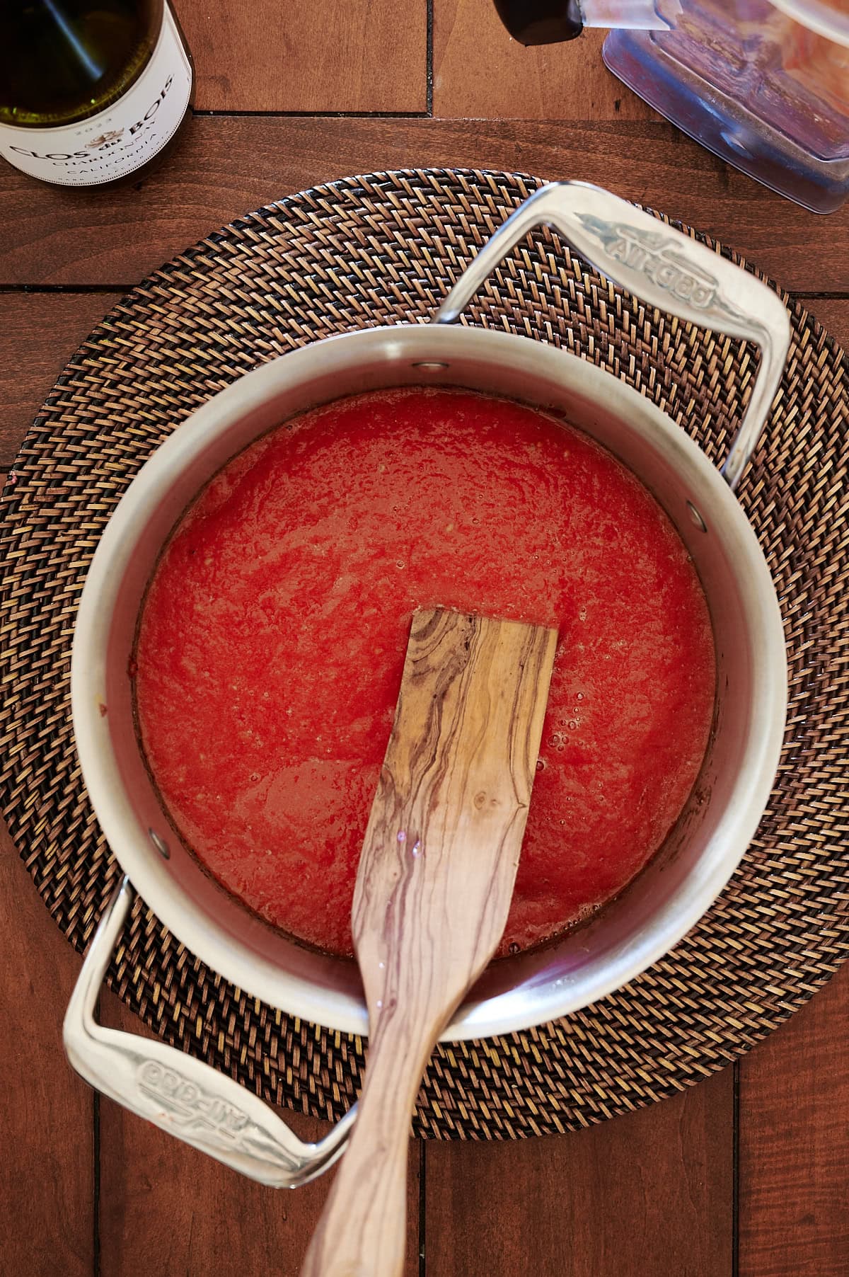 A saucepan filled with tomato sauce sits on a woven placemat, with a wooden spatula resting inside. A wine bottle is partially visible in the top left corner.